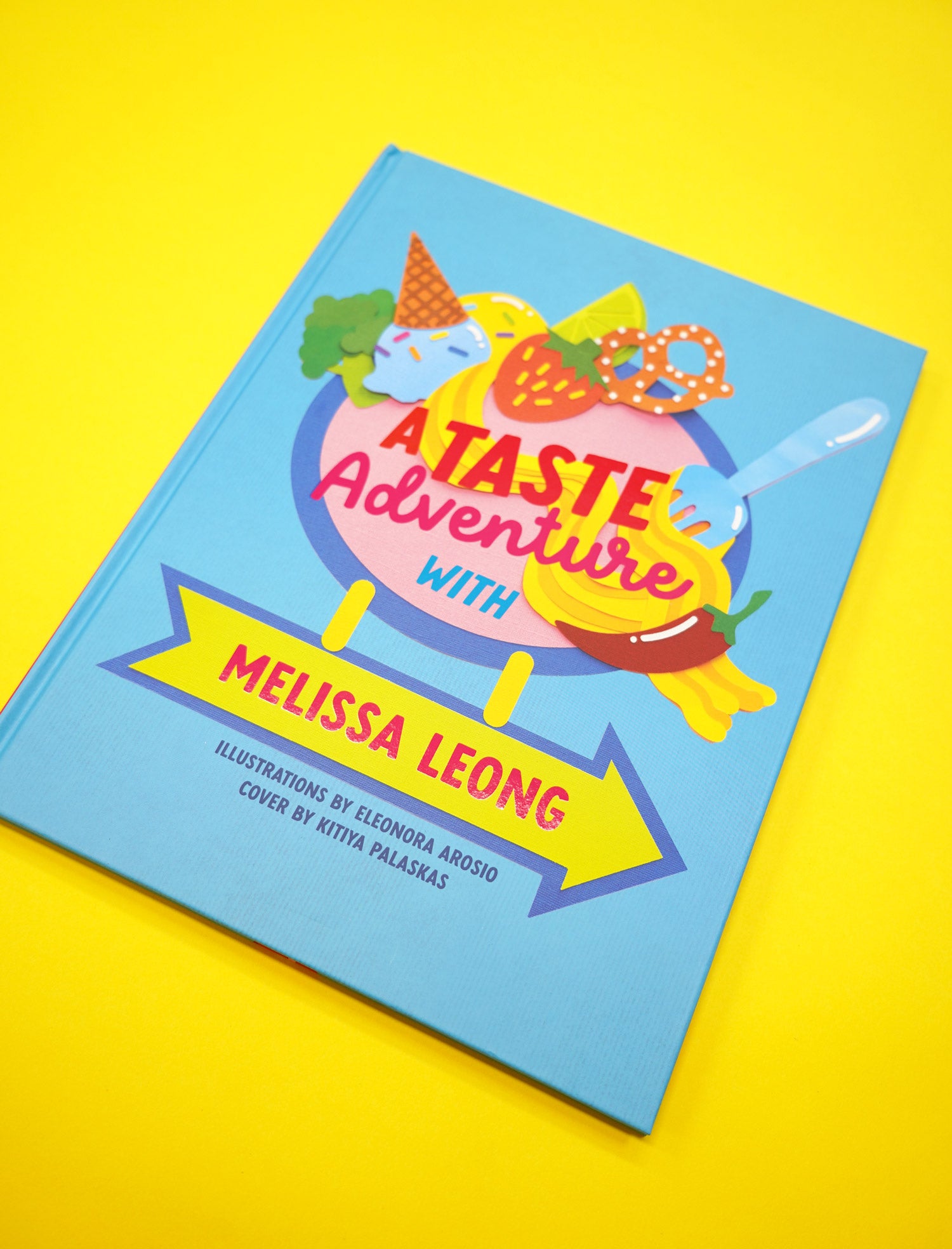 The book "A Taste Adventure" sits in a yellow background. The front cover features handmade craft based paper illustration of food by Kitiya Palaskas