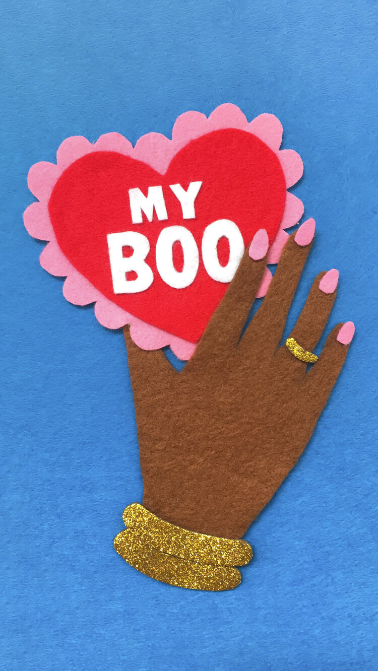 A felt collage illustration on a blue background of a brown hand holding a heart shaped valentine card that says “My boo” in white text.