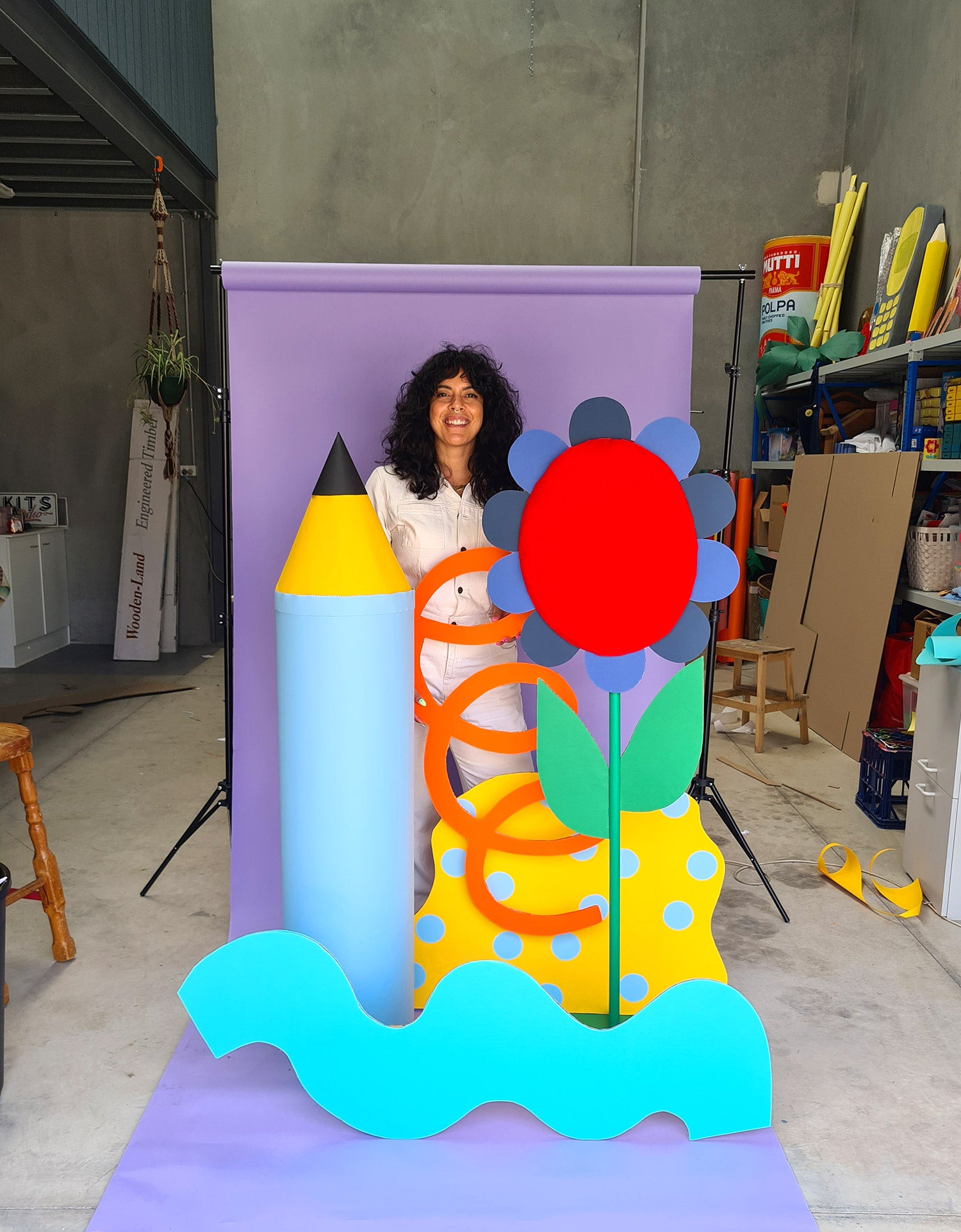 Kitiya, a woman with black hair and a white jumpsuit, stands in her design studio against a purple paper backdrop. Around her is a collection of playful, oversized cardboard props.