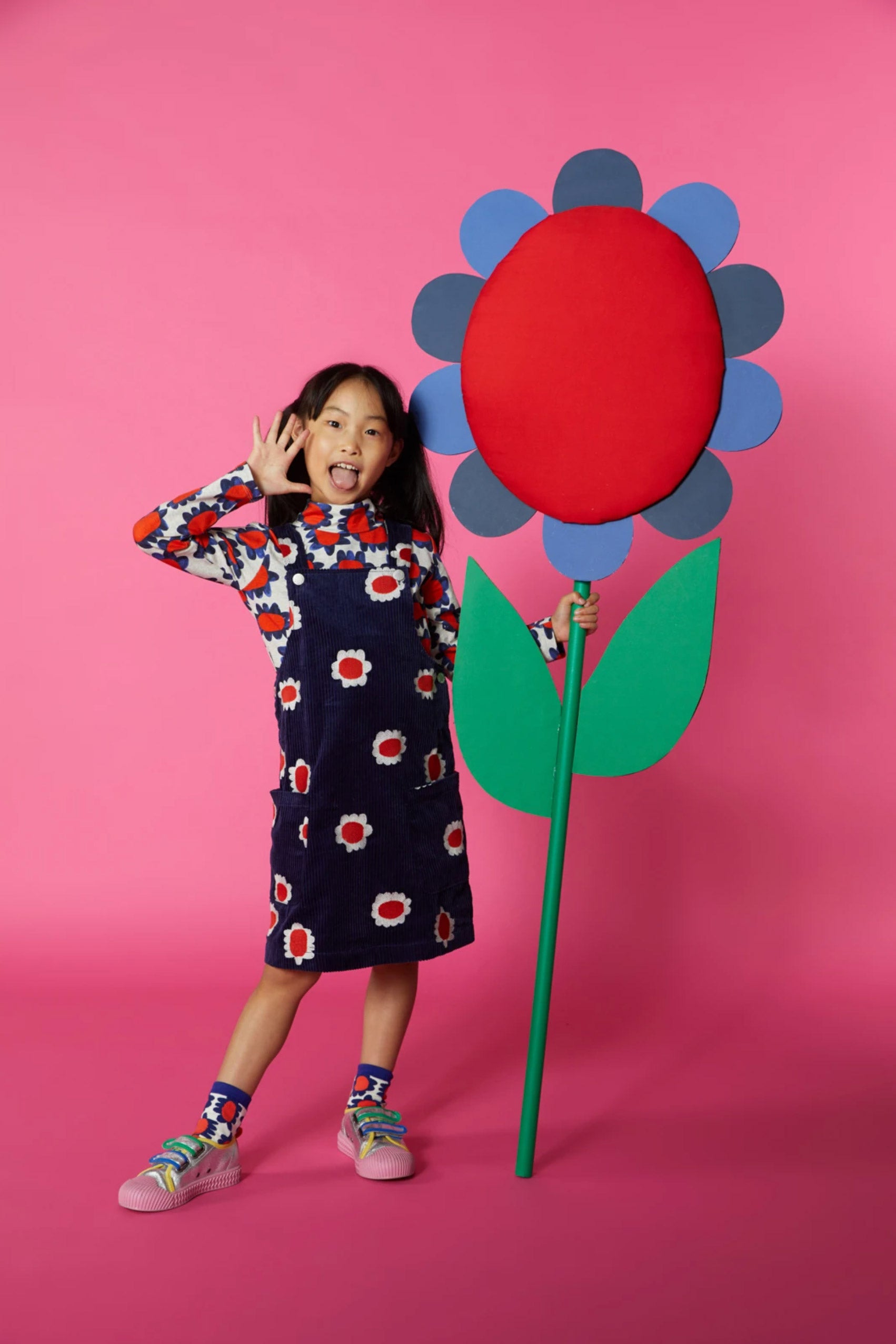 A girl in a colourful outfit stands against a dark pink background with an oversized wooden flower prop.