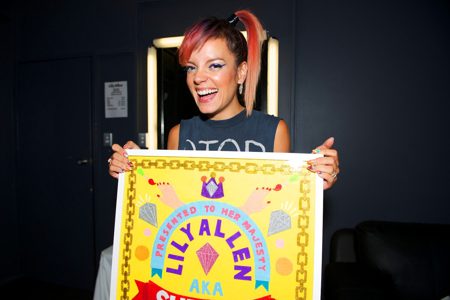 The pop singer Lily Allen smiles at the camera, holding a colourful felt collaged artwork with a yellow background, and colourful motifs and words.