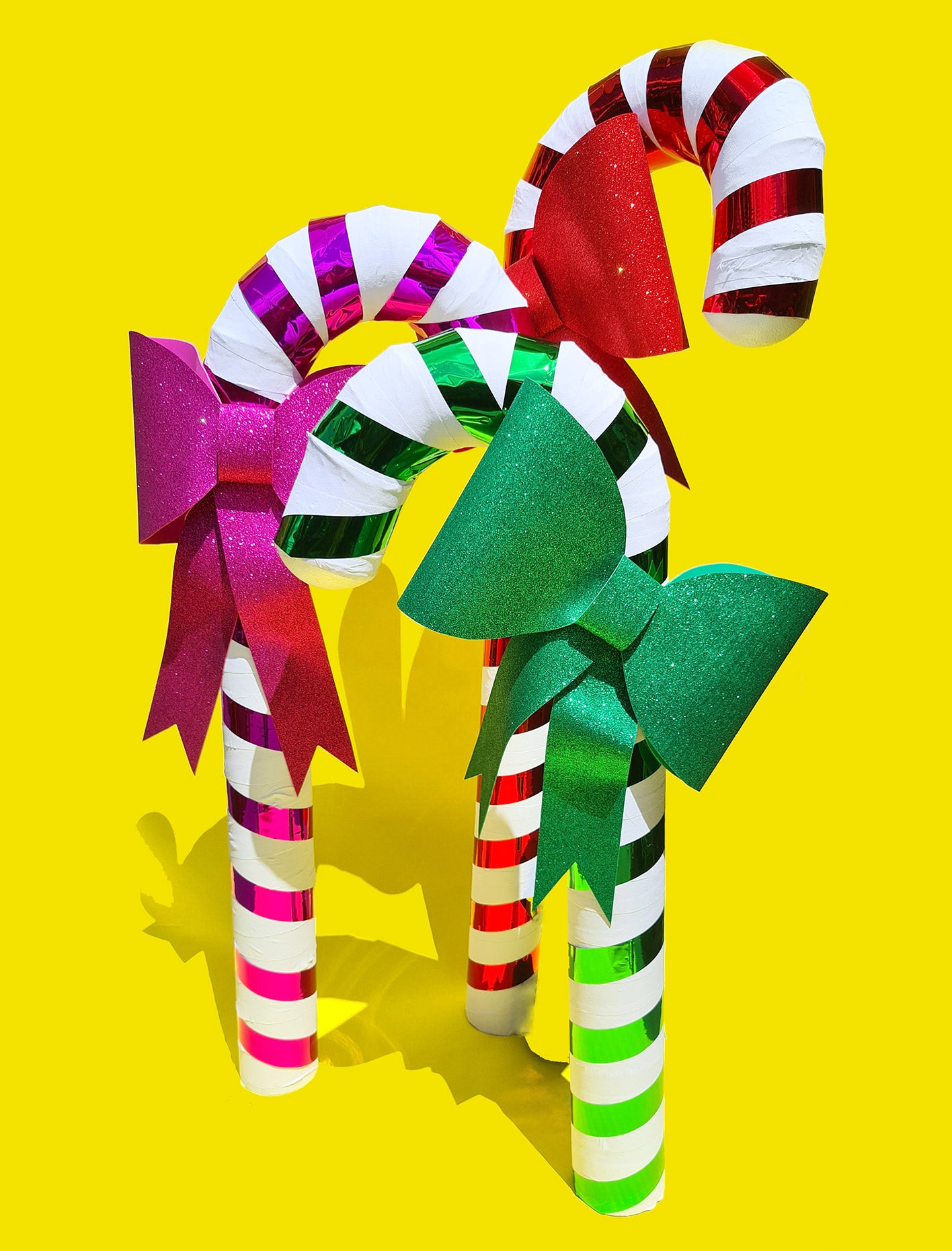 Three giant handmade candy cane Christmas props in pink, red and green sit on a yellow background, each with a matching glittery bow.