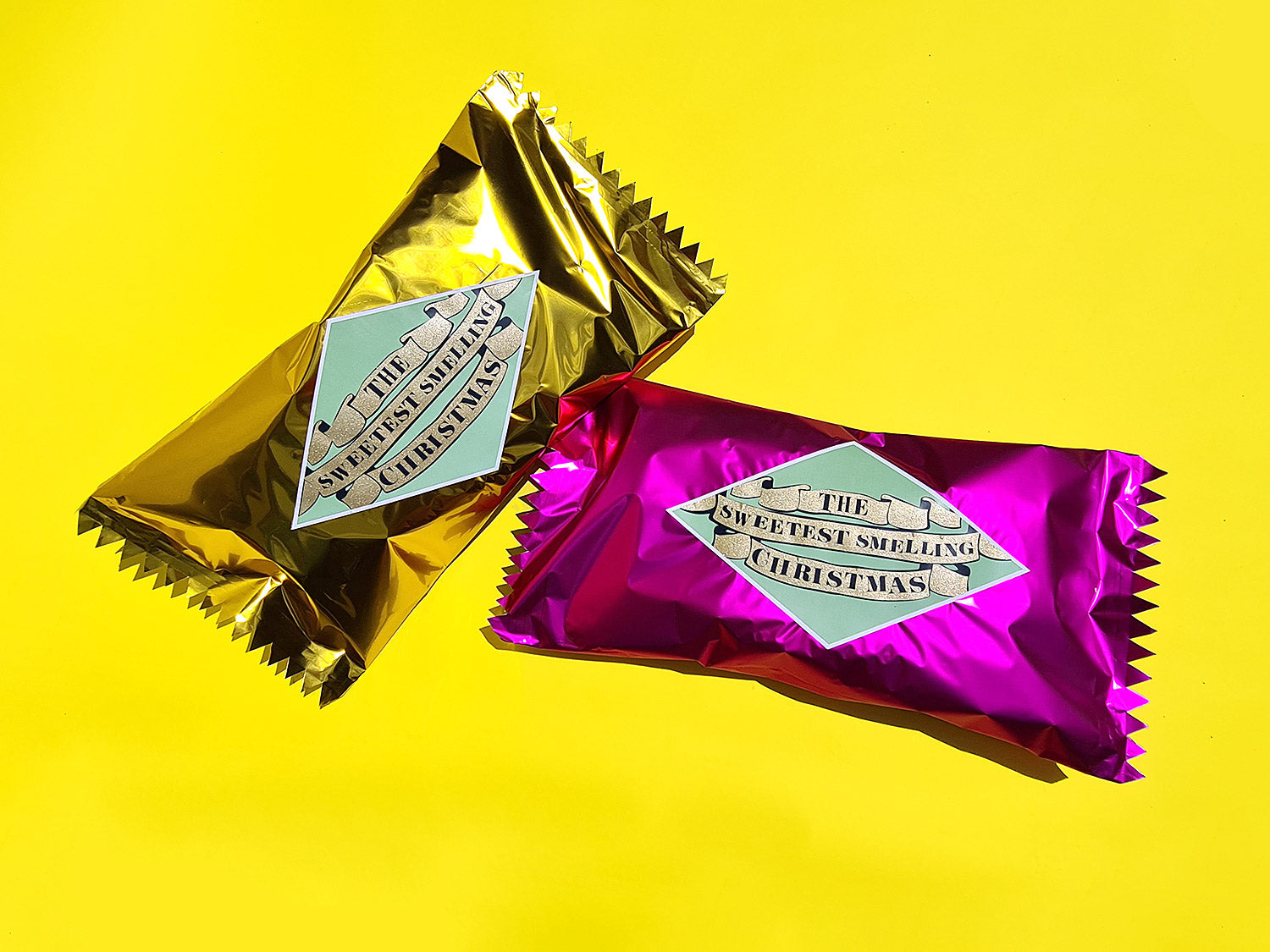 Two, giant wrapped candies in metallic pink and gold, with retro-style labels sit on a yellow background. 