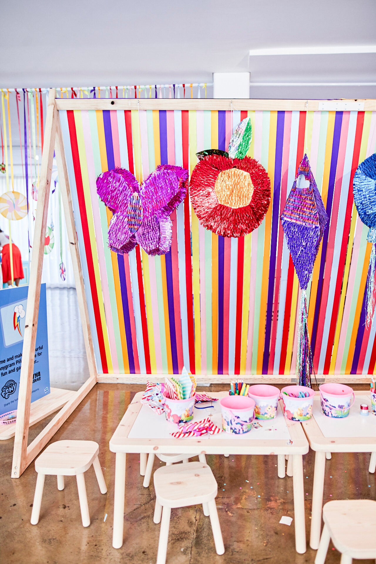 A collection of brightly coloured metallic piñatas hang against a backdrop of rainbow ribbons. In front is a long kids table covered in craft supplies. 