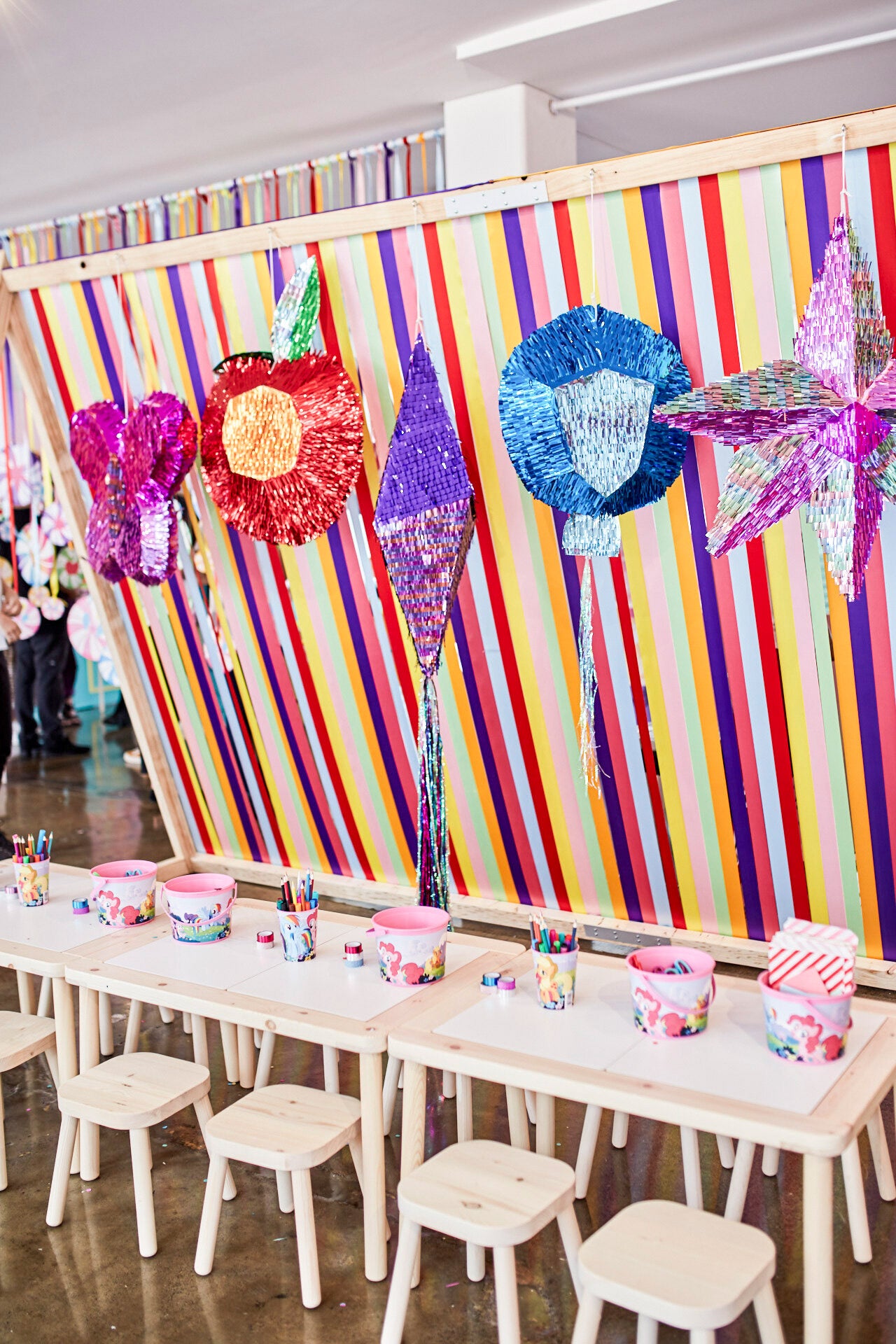 A collection of brightly coloured metallic piñatas hang against a backdrop of rainbow ribbons. Children and a giant plus My Little Pony gather in front. 