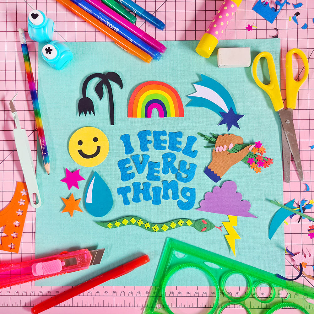 A papercraft collage illustration on a creative studio desk, depicting a collection of colourful motifs and the words “I feel everything” in black text. The artwork is surrounded by craft supplies and stationery.