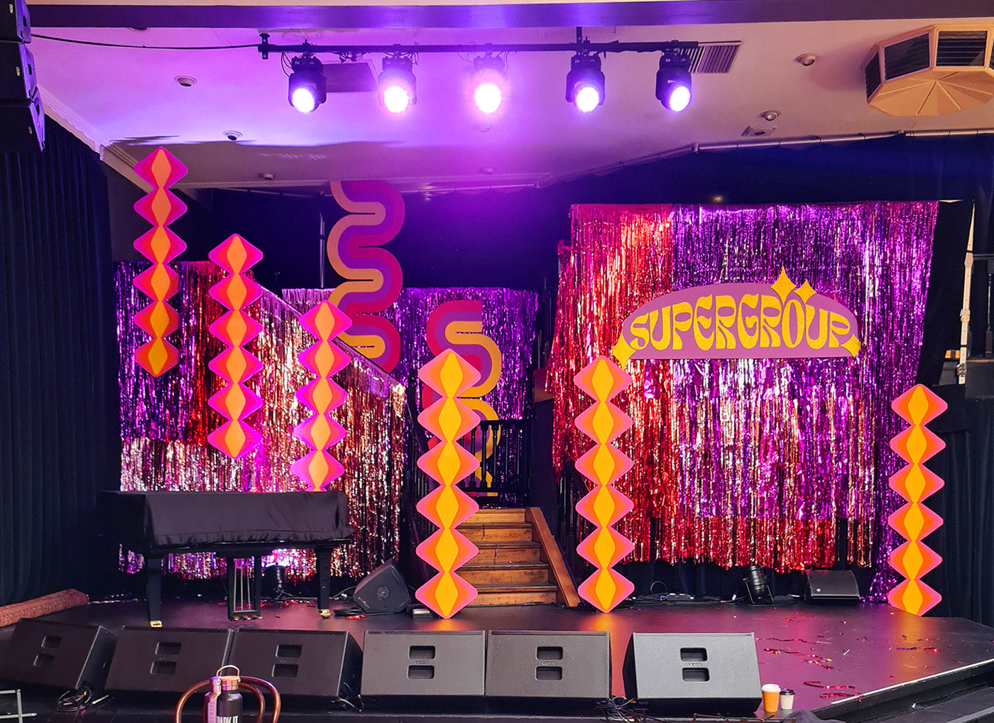 A live music venue featuring a colourful stage set design with shimmer curtains and retro-inspired cardboard cut-out set pieces. 