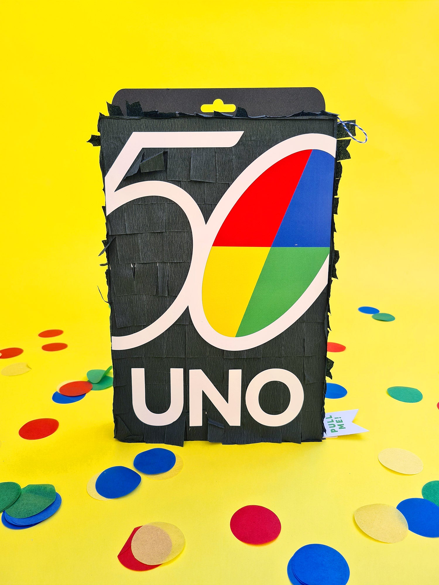 A collection of piñatas in the shape of black UNO playing card boxes, with the UNO logo on the front sit on a yellow background, surrounded by confetti in blue, yellow, red and green. 