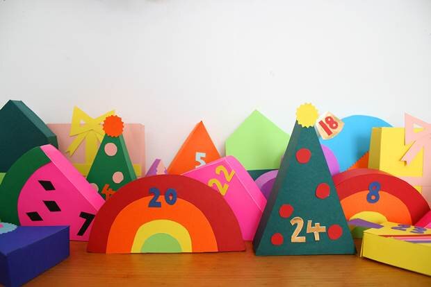 A collection of handmade colourful papercraft boxes in a variety of shapes sit on a wooden shelf. Each box is numbered to form a colourful advent calendar.