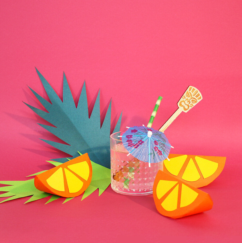 A tropical cocktail in a textured glass sits among a collection of tropical papercraft props, against a pink background.