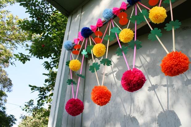 A colourful pom-pom and paper straw garland hangs in the doorway of a rustic building. The garland is handmade and features paper cutout shapes. 