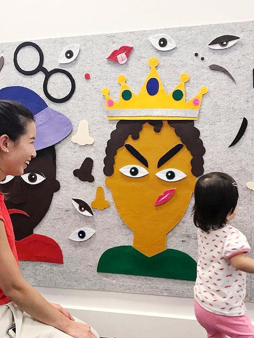 A grey felt collage installation wall with various colourful facial features and accessories stuck to it. A mother and child sit in front of the wall.