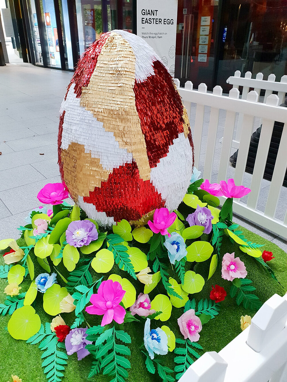 A giant red, gold and white piñata egg sits in a garden of colourful paper plants and flowers, surrounded by a white picket fence, inside a shopping centre.