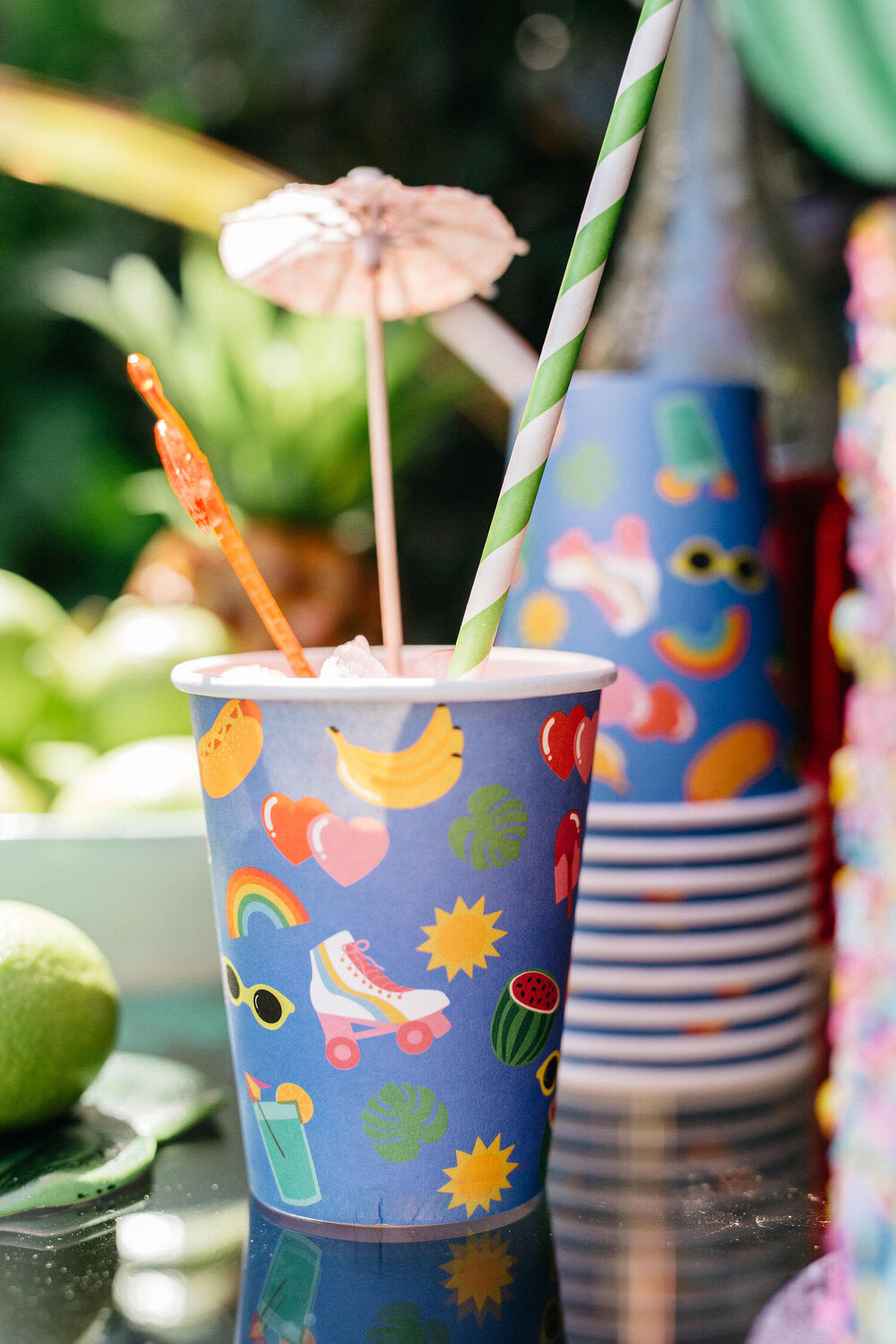 A collection of vibrant partyware in a party setting. The partyware includes plates, cups, party bags and piñatas and features a blue background, with tropical illustrated motifs. 