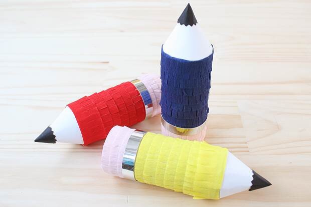 Three handmade mini pencil piñatas in blue, yellow and red sit on a wooden desk.