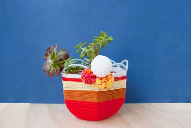 A colourful upcycled lampshade has been transformed into a yarn woven planter basket. It sits on a wooden table against a blue wall and is decorated with pom poms and filled with succulents.