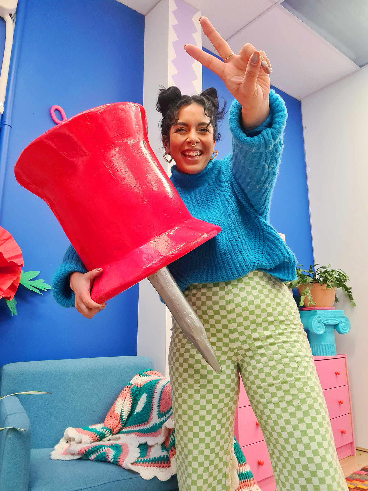  Kitiya, a woman of colour with black hair, a blue sweater and green checkered pants stands in her colourful art studio holding a giant, oversized red papier mâché pin/thumbtack prop, and giving a peace sign