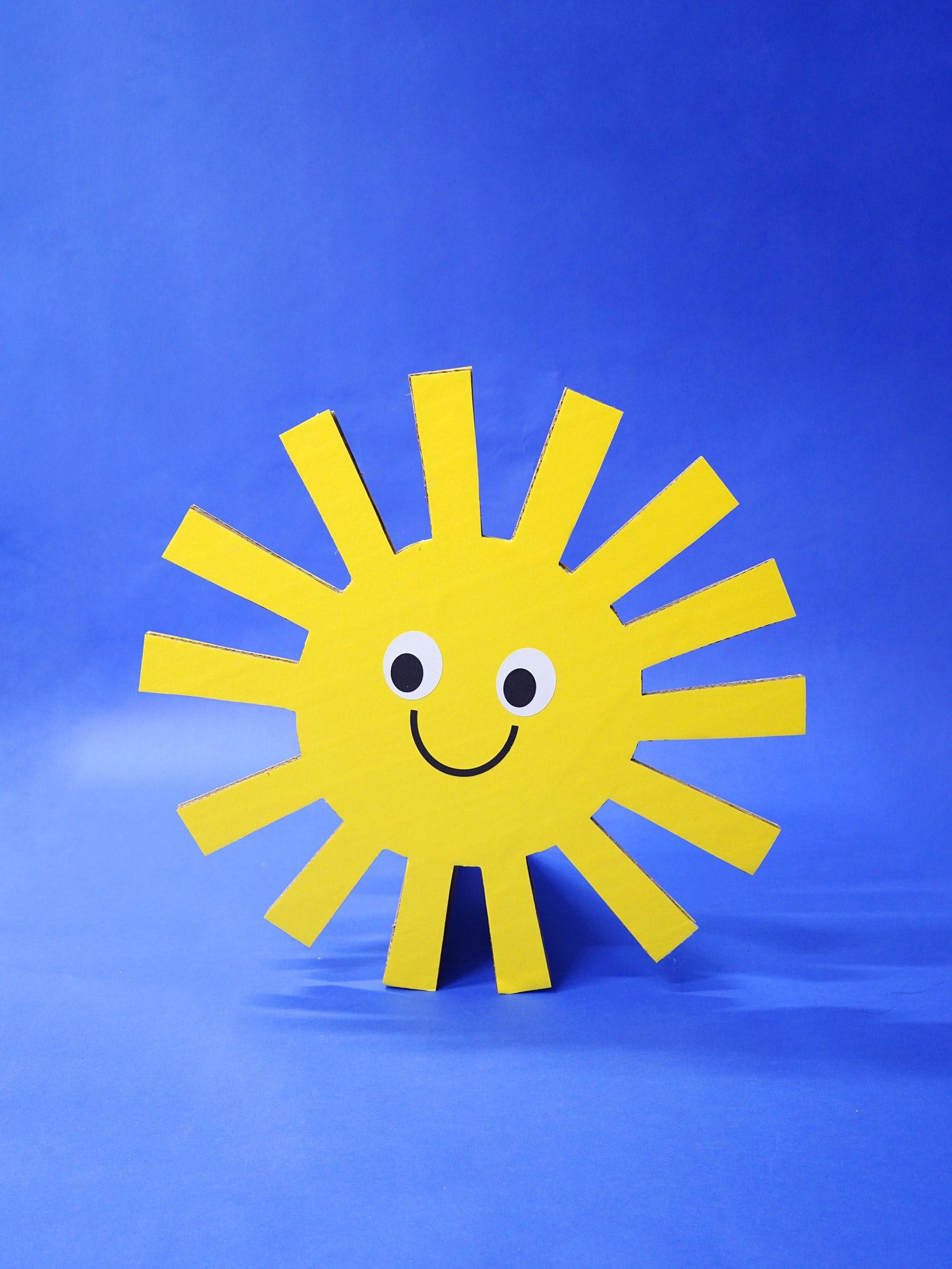 An oversized yellow cardboard cutout sun prop with a smiley face stands against a blue backdrop.
