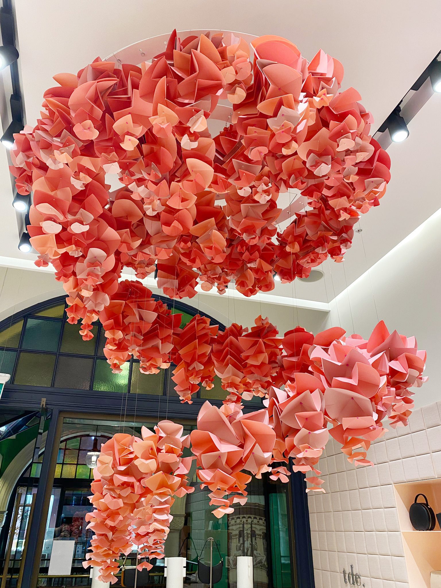 A large, pink, spiral-shaped papercraft petal installation hangs from the ceiling of a brightly lit retail store.