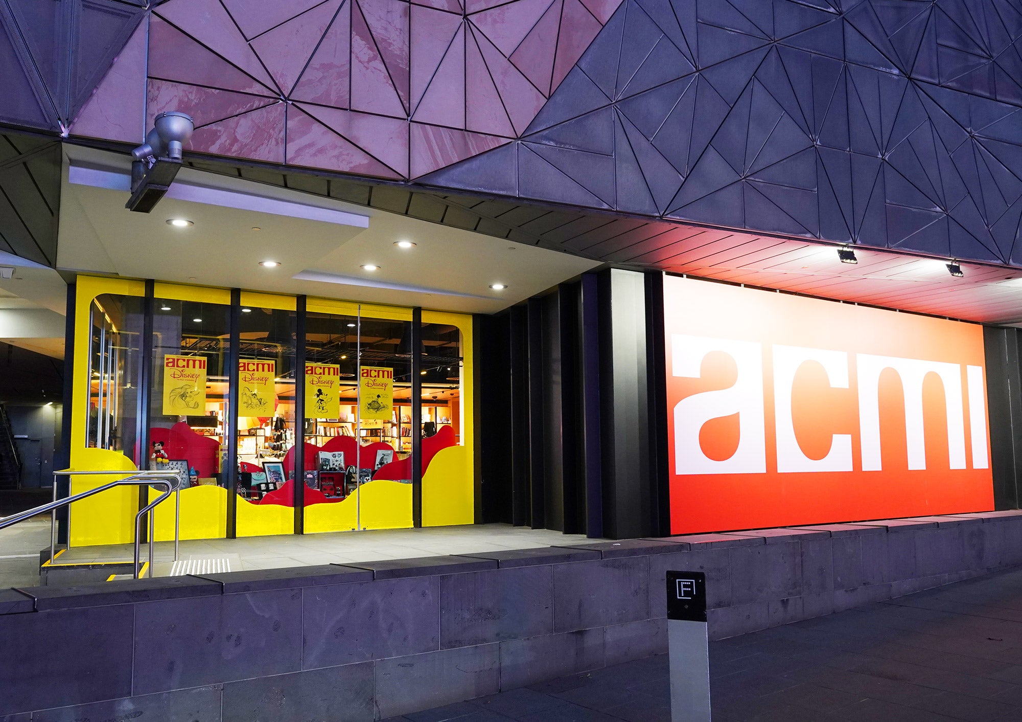 A Disney-themed retail window installation in the ACMI design shop, designed by Kitiya Palaskas Studio, with large red and white abstract window details, red abstract flat set pieces, and lots of Disney products. 