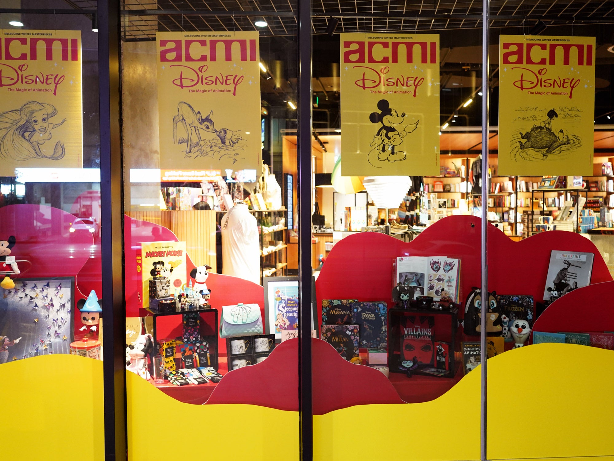 A Disney-themed retail window installation in the ACMI design shop, designed by Kitiya Palaskas Studio, with large red and white abstract window details, red abstract flat set pieces, and lots of Disney products. 