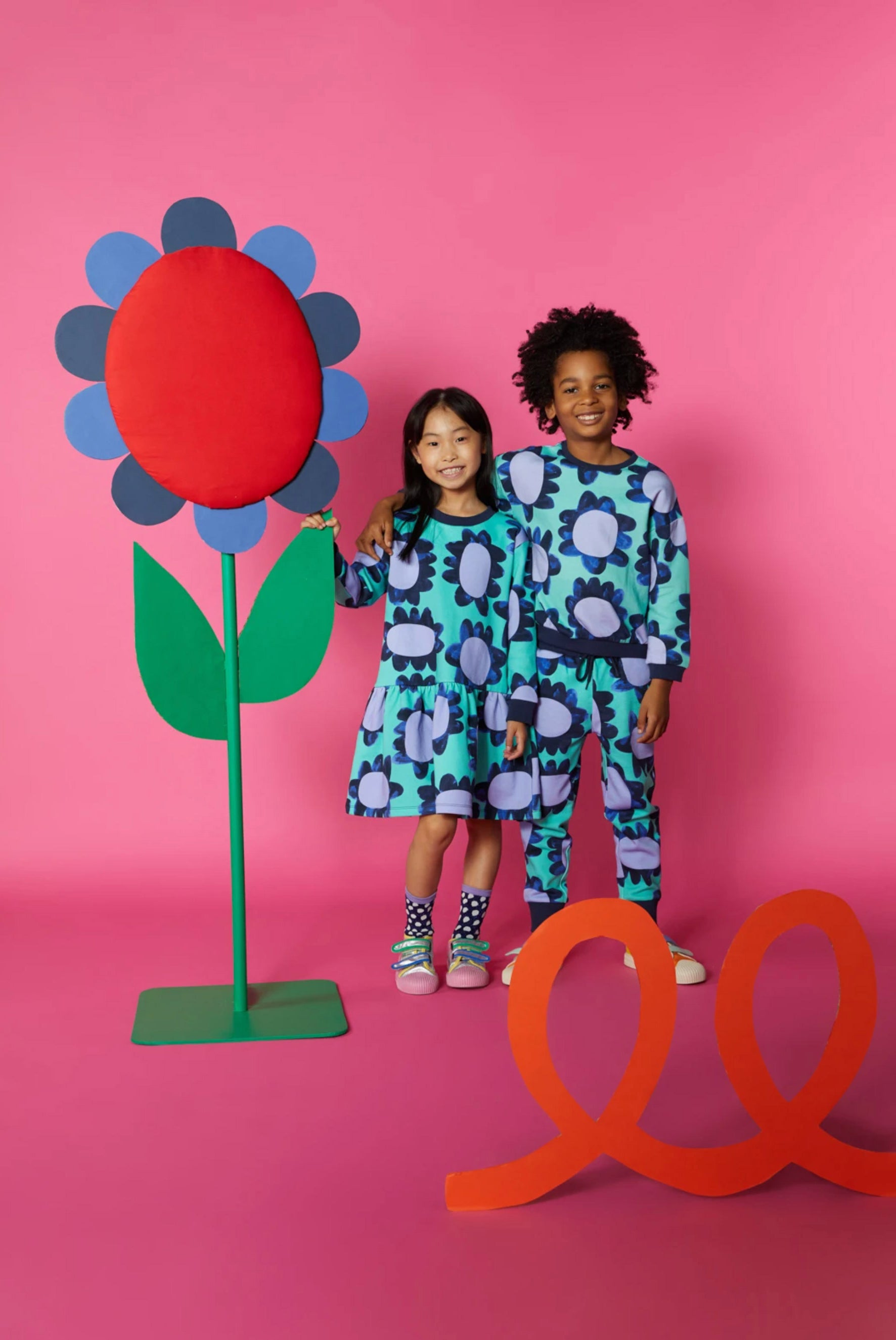 Two children in cololurful clothes stand against a dark pink background with an oversized wooden flower prop and an orange loopy cardboard cutout prop.