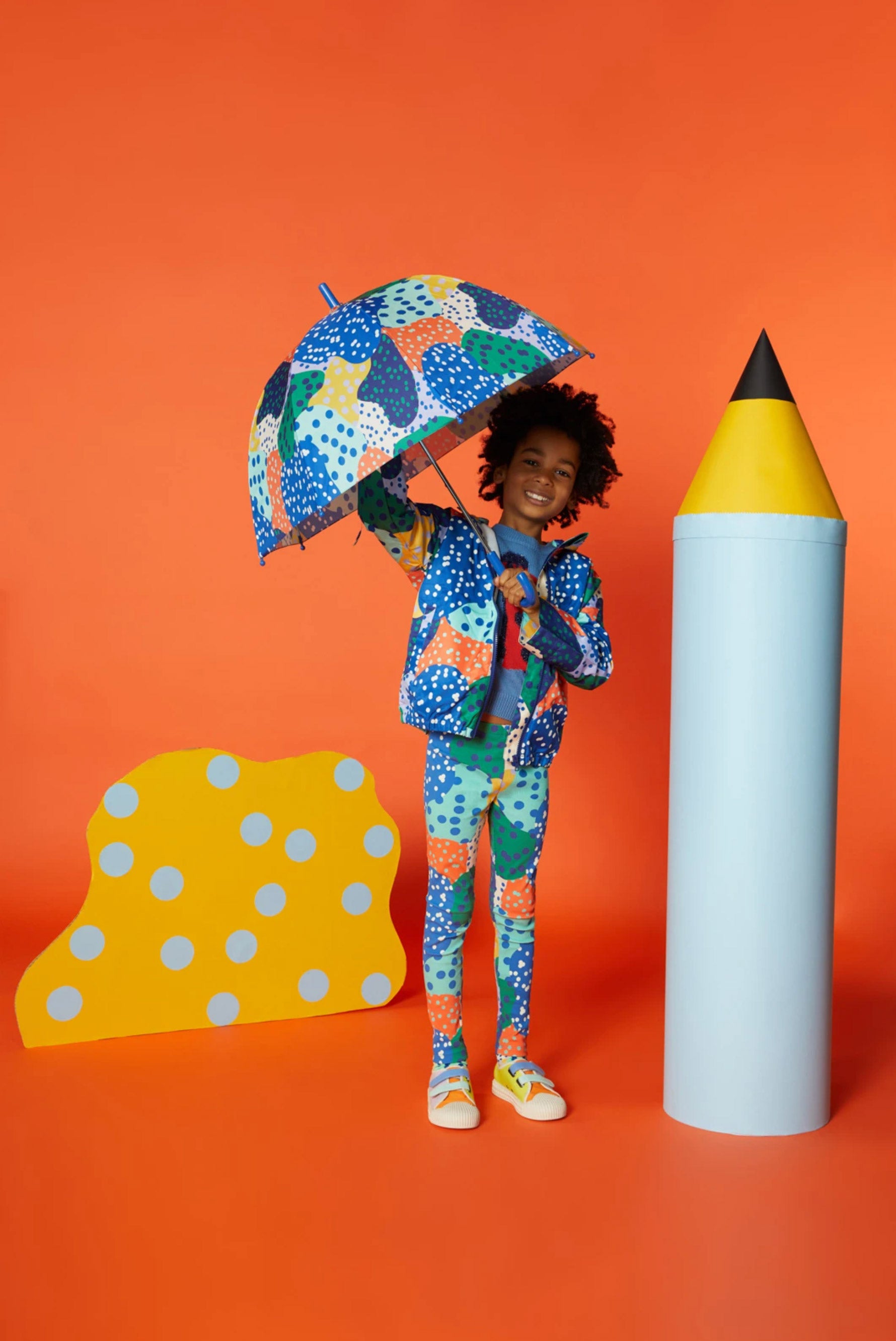 A boy with a colourful umbrella stands against an orange background with an oversized blue pencil prop, and a yellow abstract cardboard cutout prop with blue polkadots. 