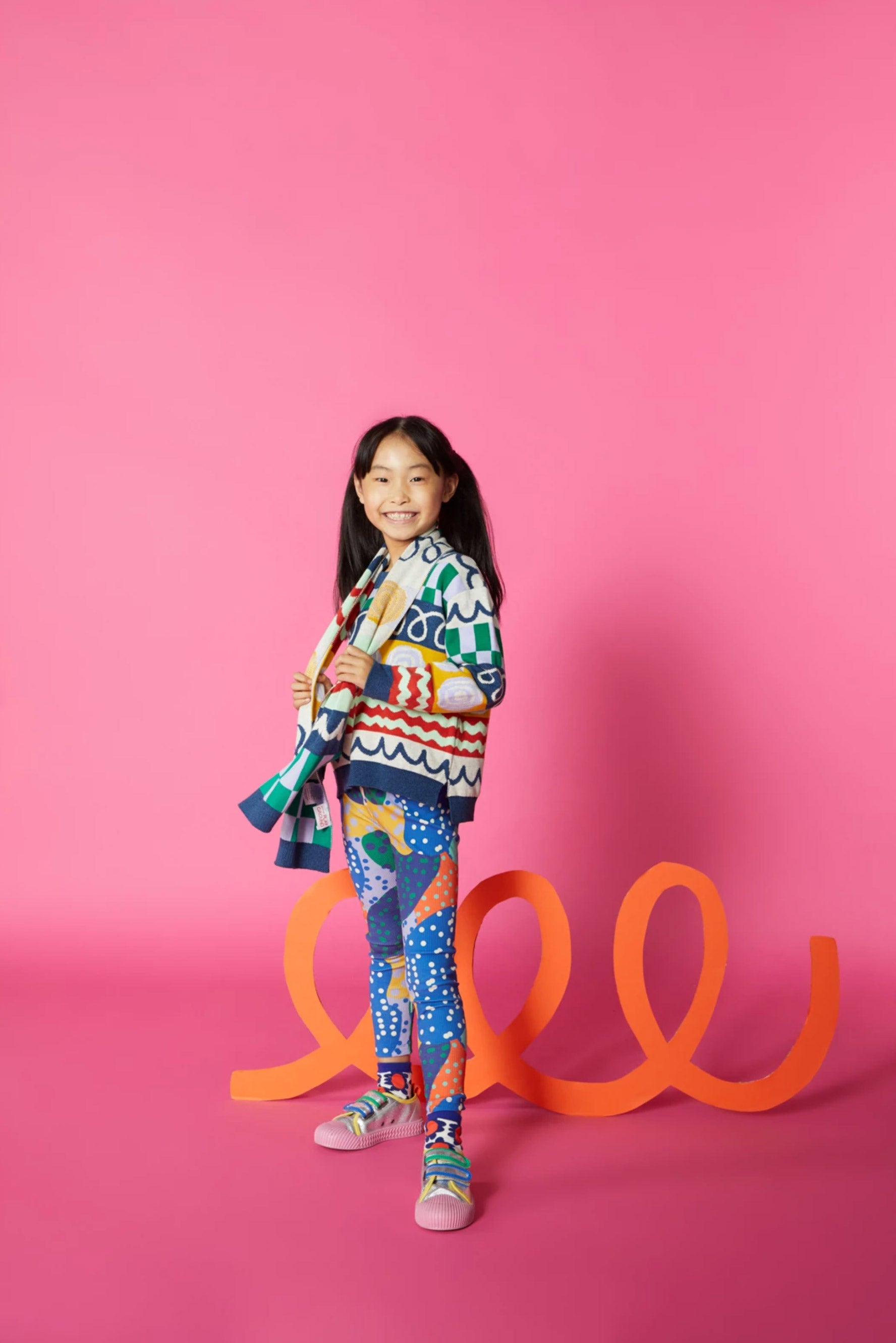 A girl in a colourful outfit stands against a dark pink background with an orange loopy cardboard cutoutprop.