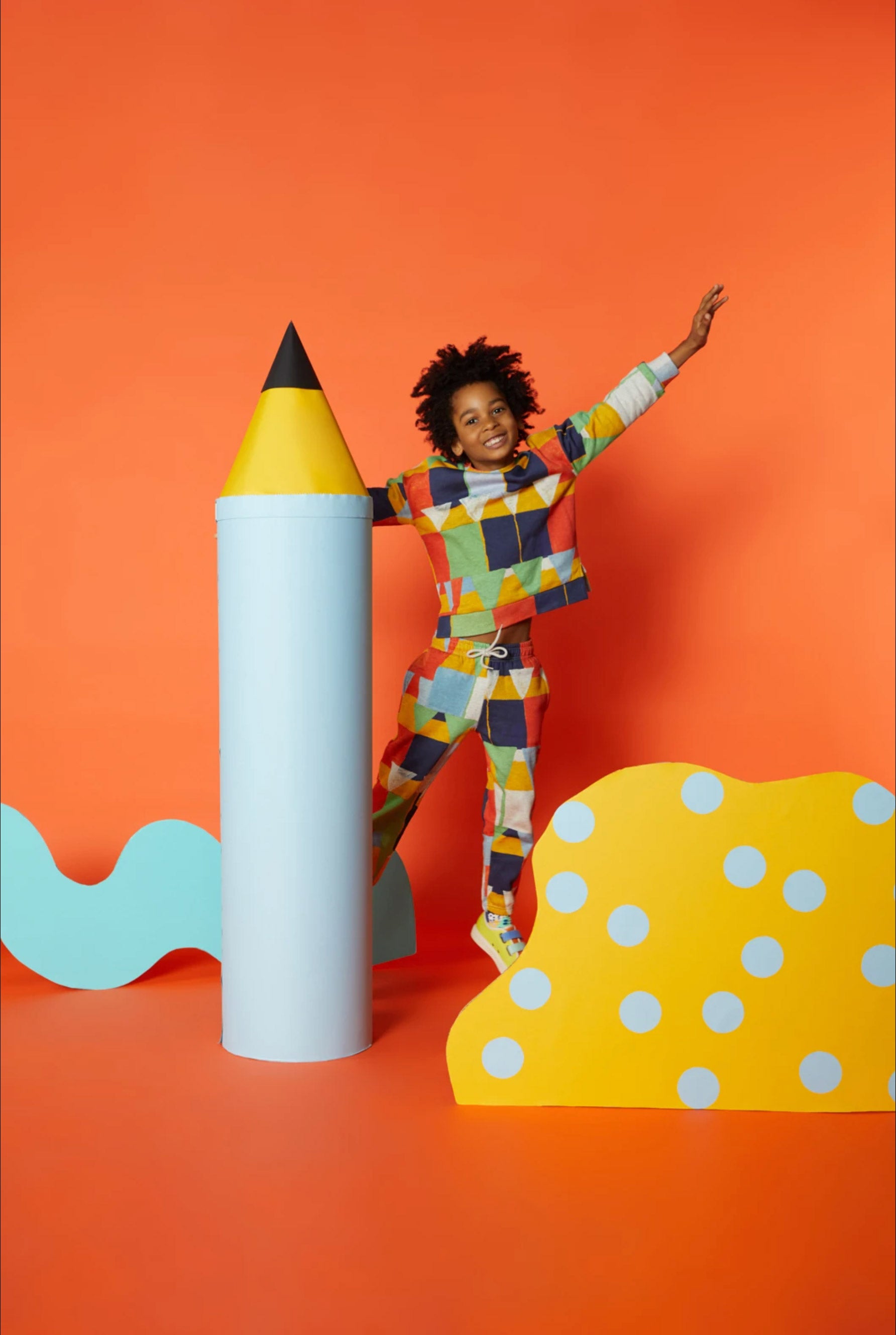 A boy in a colourful outfit jumps in front of an orange background, surrounded by a giant blue pencil prop,  wavy blue cardboard cutout prop and a yellow abstract cardboard cutout prop with blue polkadots. 