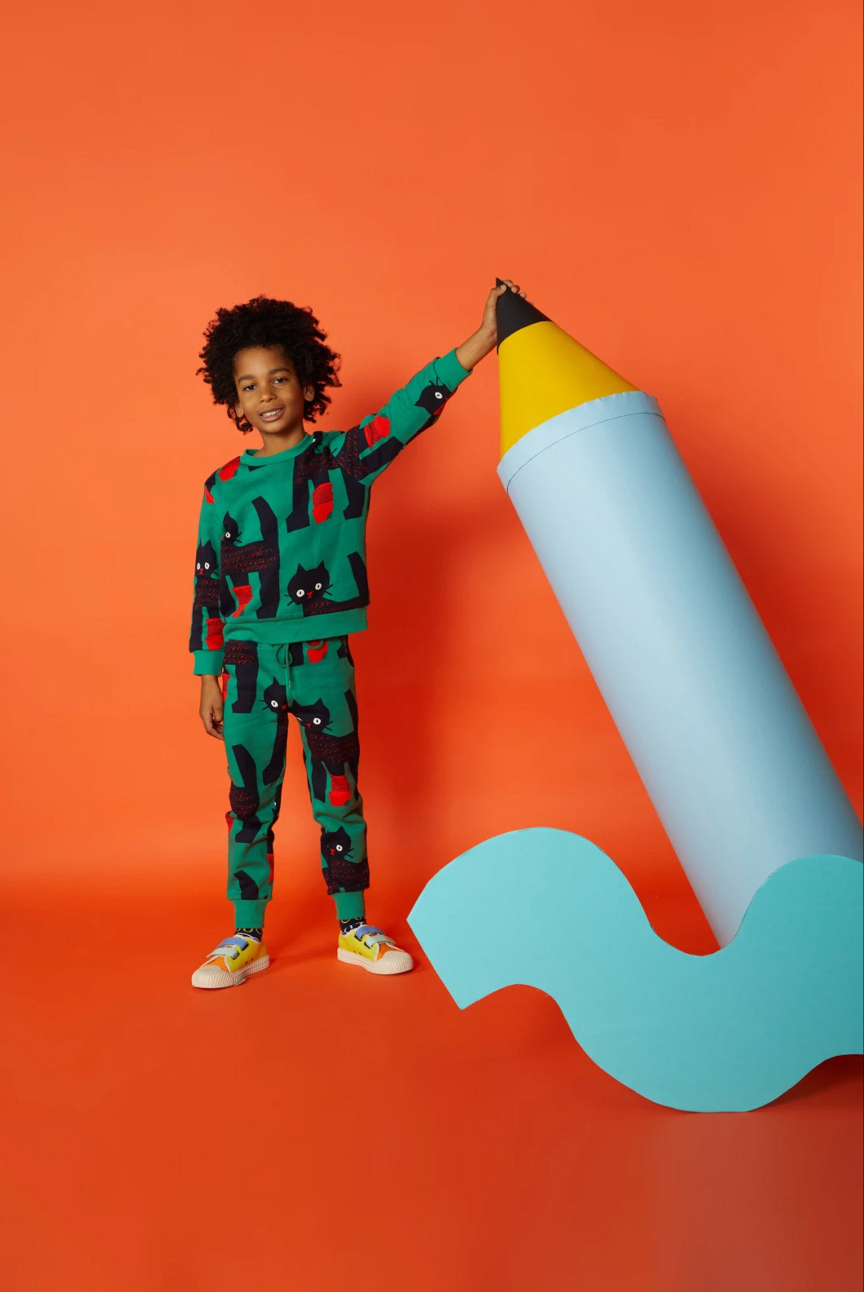 A boy in colourful clothes stands against an orange background with an oversized blue pencil prop, and a yellow abstract cardboard cutout prop with blue polkadots. 