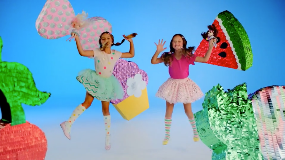 Two young girls holding dolls jump in mid air, surrounded by a collection of giant, shiny, handmade props in the shapes of a pineapple, bow, cherries, cupcake and watermelon.