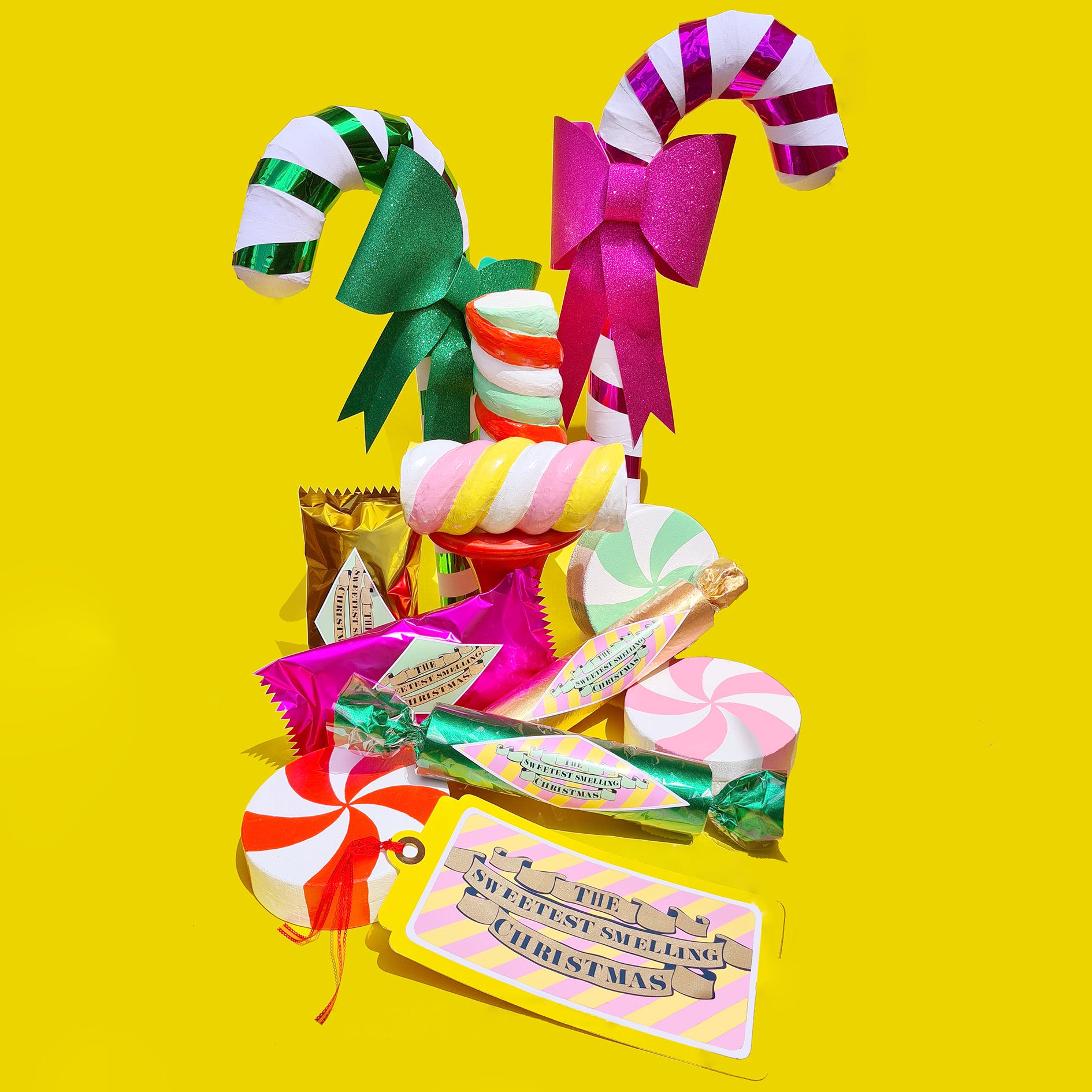A collection of bright, shiny, giant, handmade Christmas candy props sit together on a yellow background. There are candy canes, wrapped lollies, marshmallow twists, and peppermint swirls.