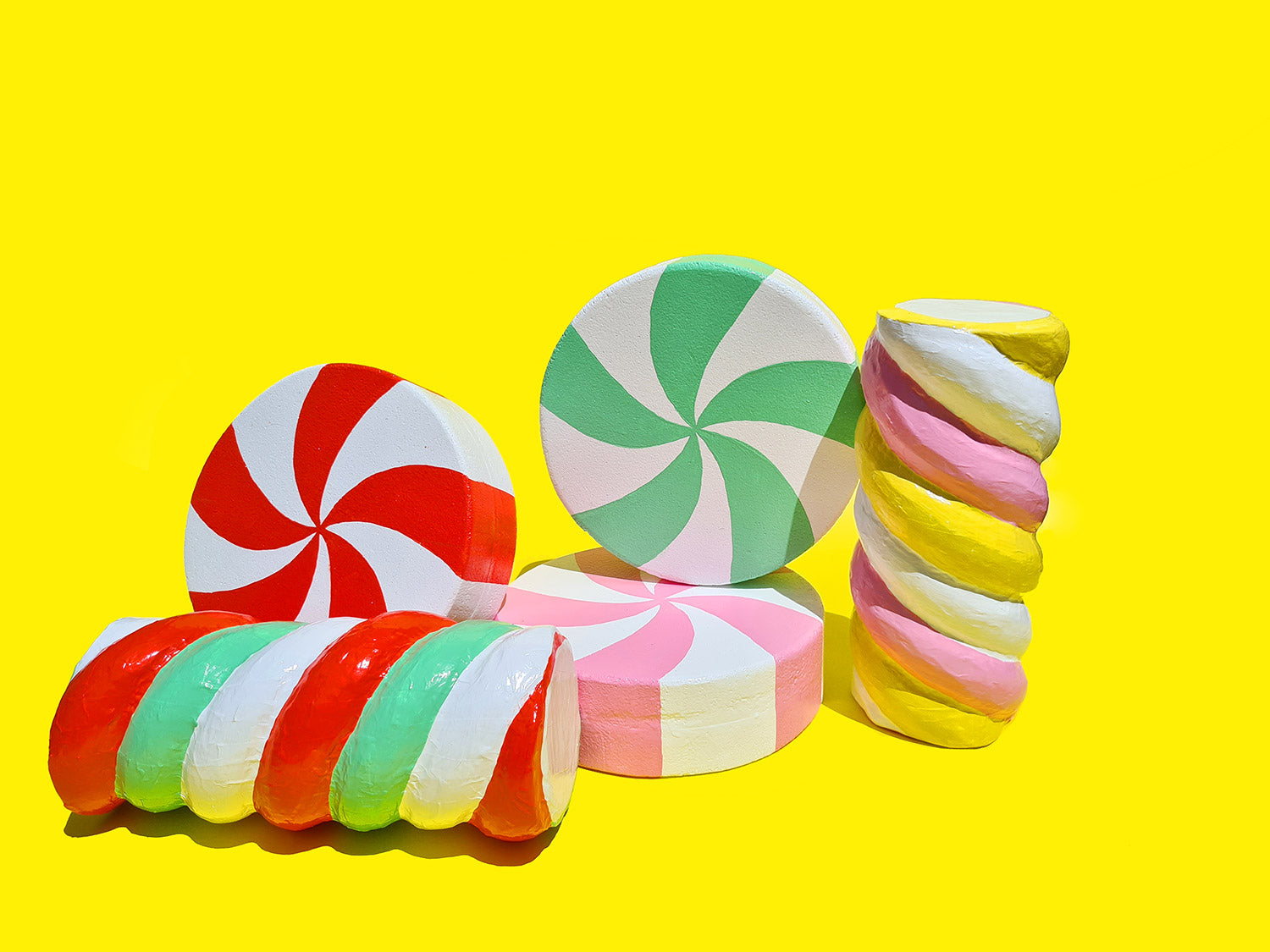 A collection of giant, handmade Christmas candy props sit together on a yellow background, including marshmallow twists and peppermint swirls. 