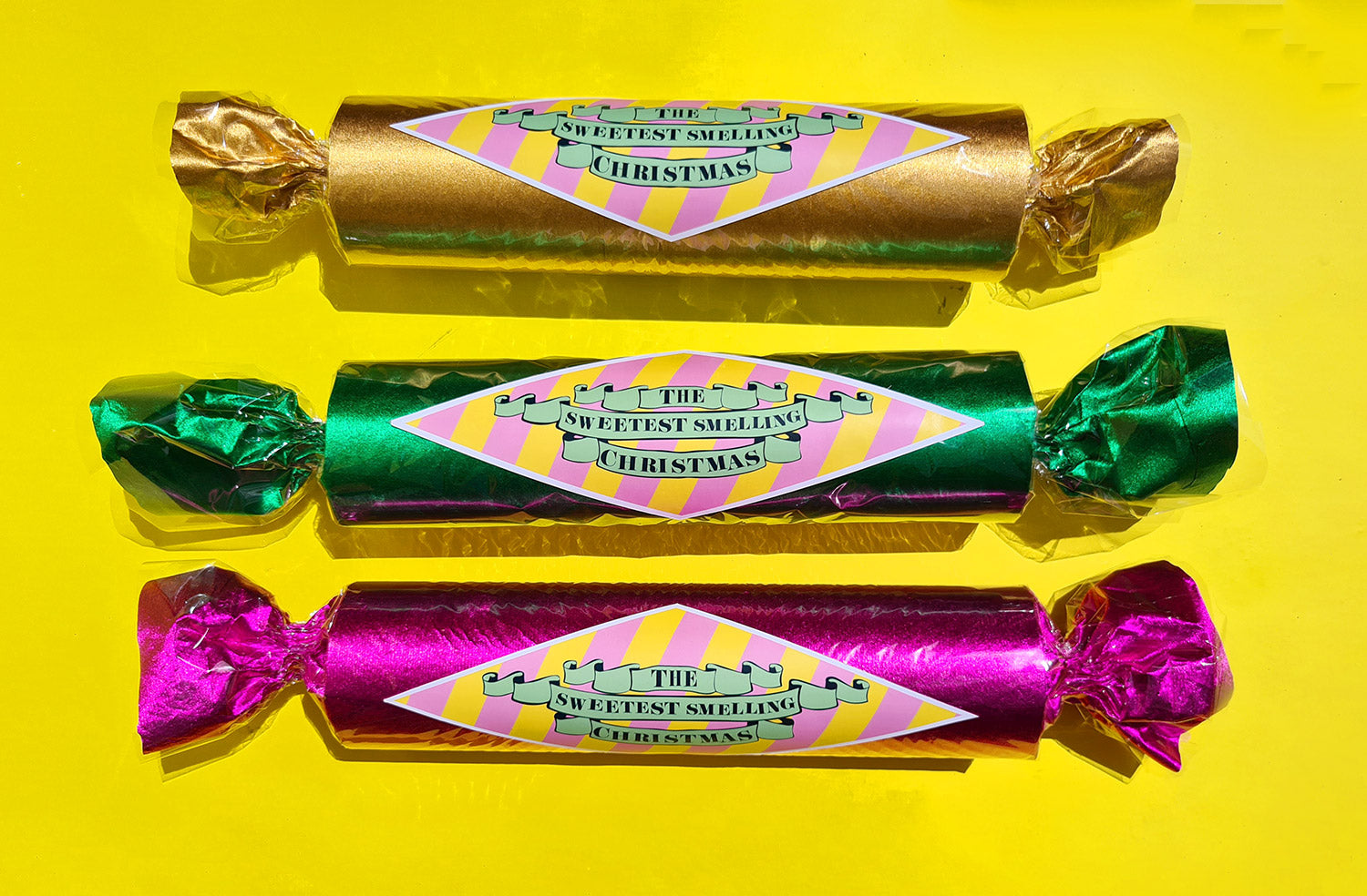 Three, giant wrapped candies in metallic pink, gold and green, with retro-style labels sit on a yellow background. 