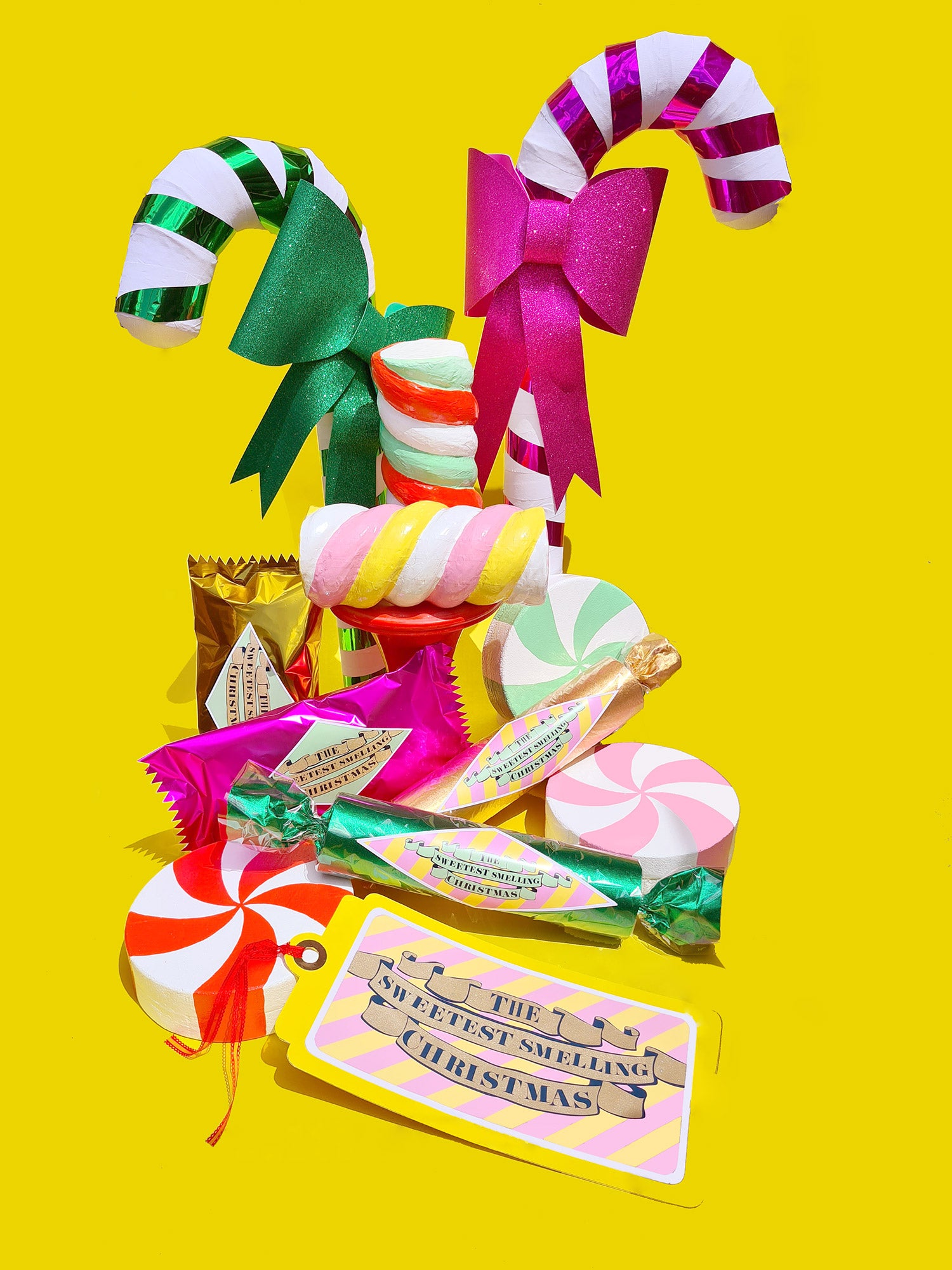 A collection of bright, shiny, giant, handmade Christmas candy props sit together on a yellow background. There are candy canes, wrapped lollies, marshmallow twists, and peppermint swirls. 