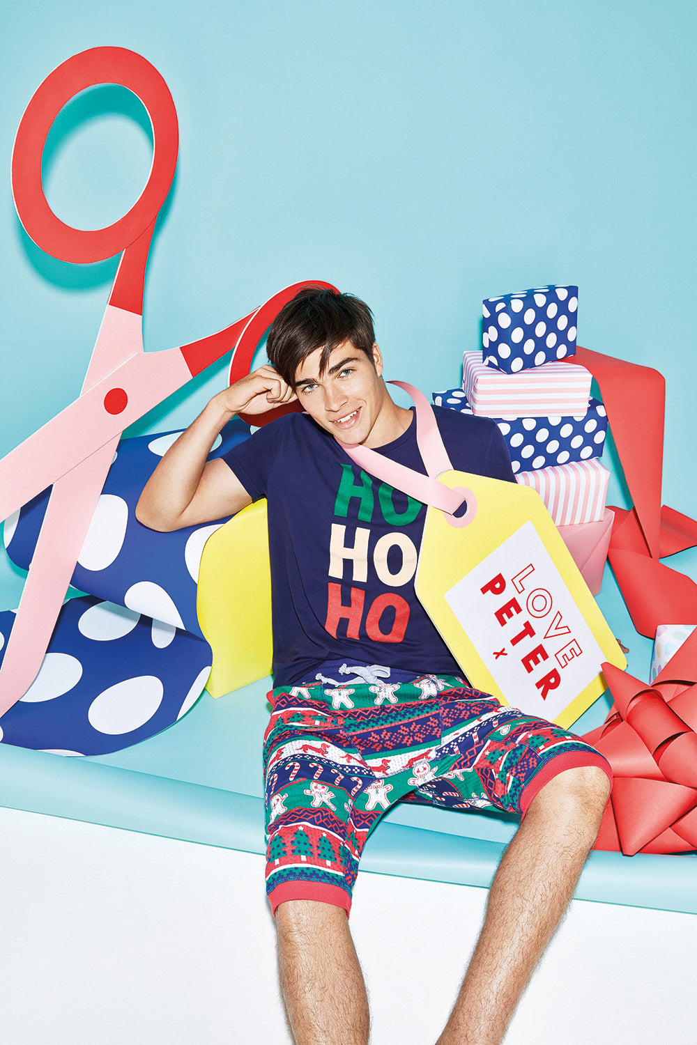 A man in Christmas-themed pajamas sits on a blue backdrop, surrounded by colourful present props, a giant handmade red and pink papercraft scissors prop, and a giant yellow handmade papercraft gift tag. 