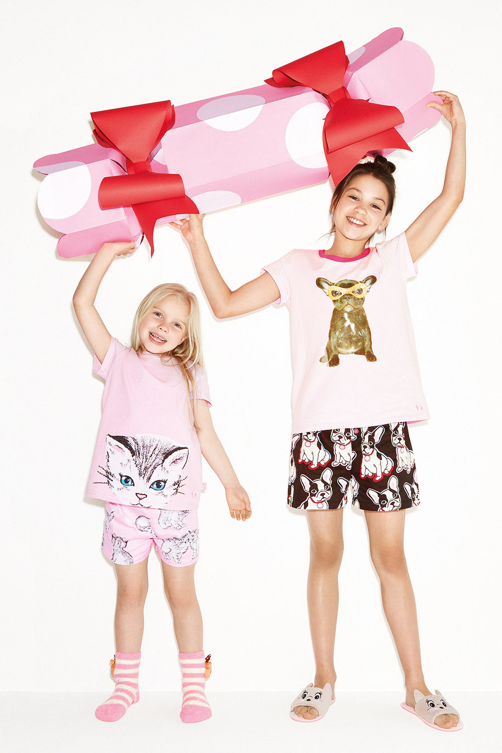 Two young girls in Christmas-themed pajamas stand in front of a yellow backdrop holding a giant handmade pink Christmas bon bon prop with red bows. 