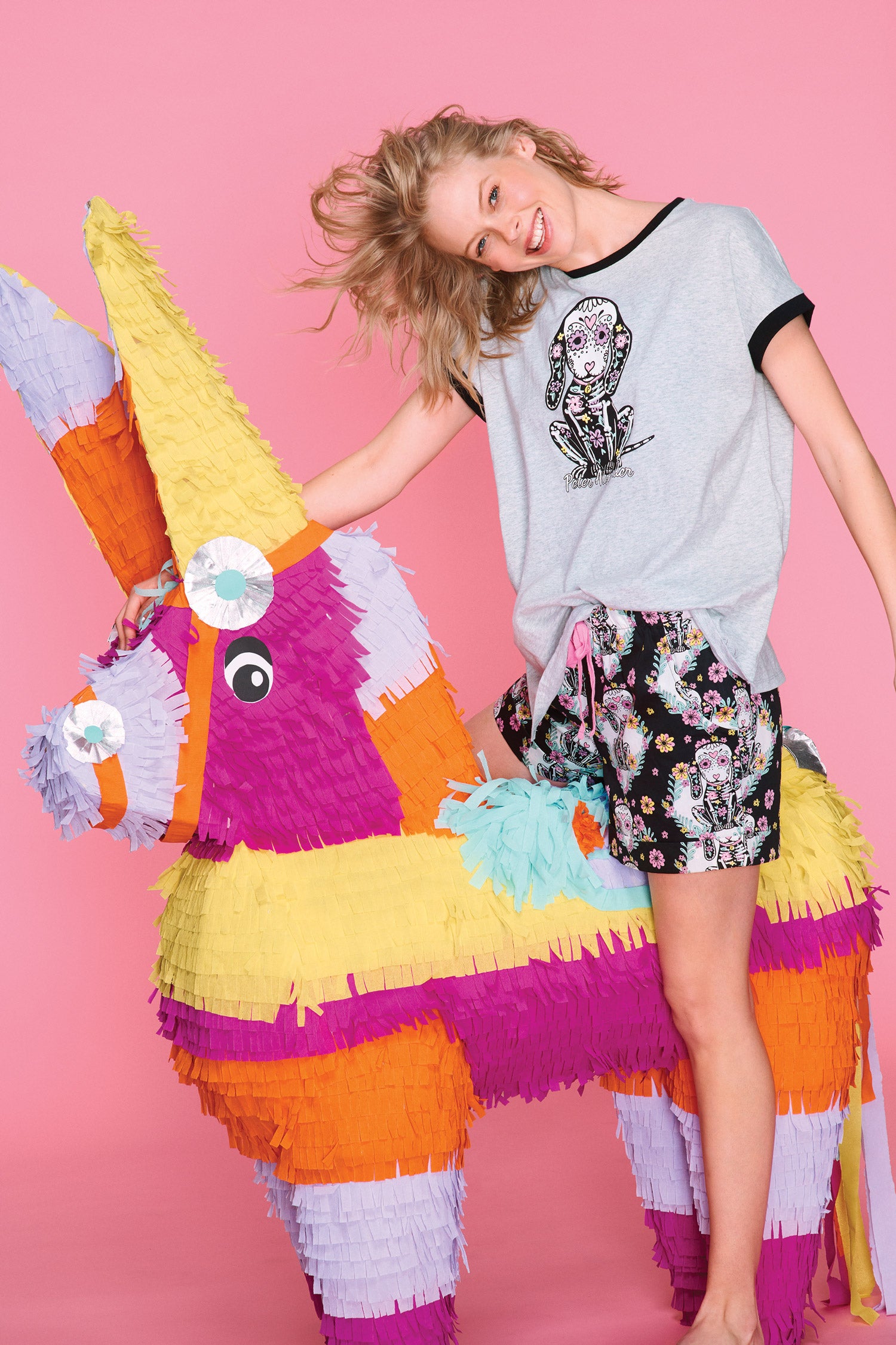A giant, colorful, oversized, handmade donkey piñata stands against a pink background. A woman in piñata-themed pajamas is riding the donkey.
