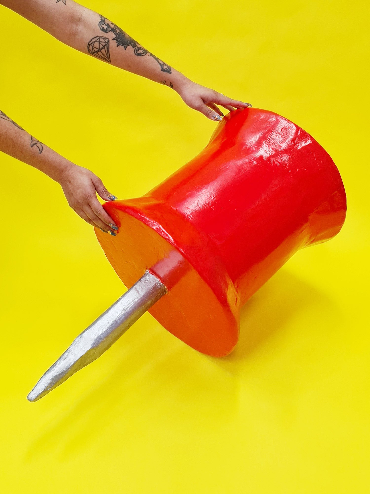 A pair of arms with tattoos hold a giant, handmade, oversized red papier-mâché pin prop against a yellow background. 
