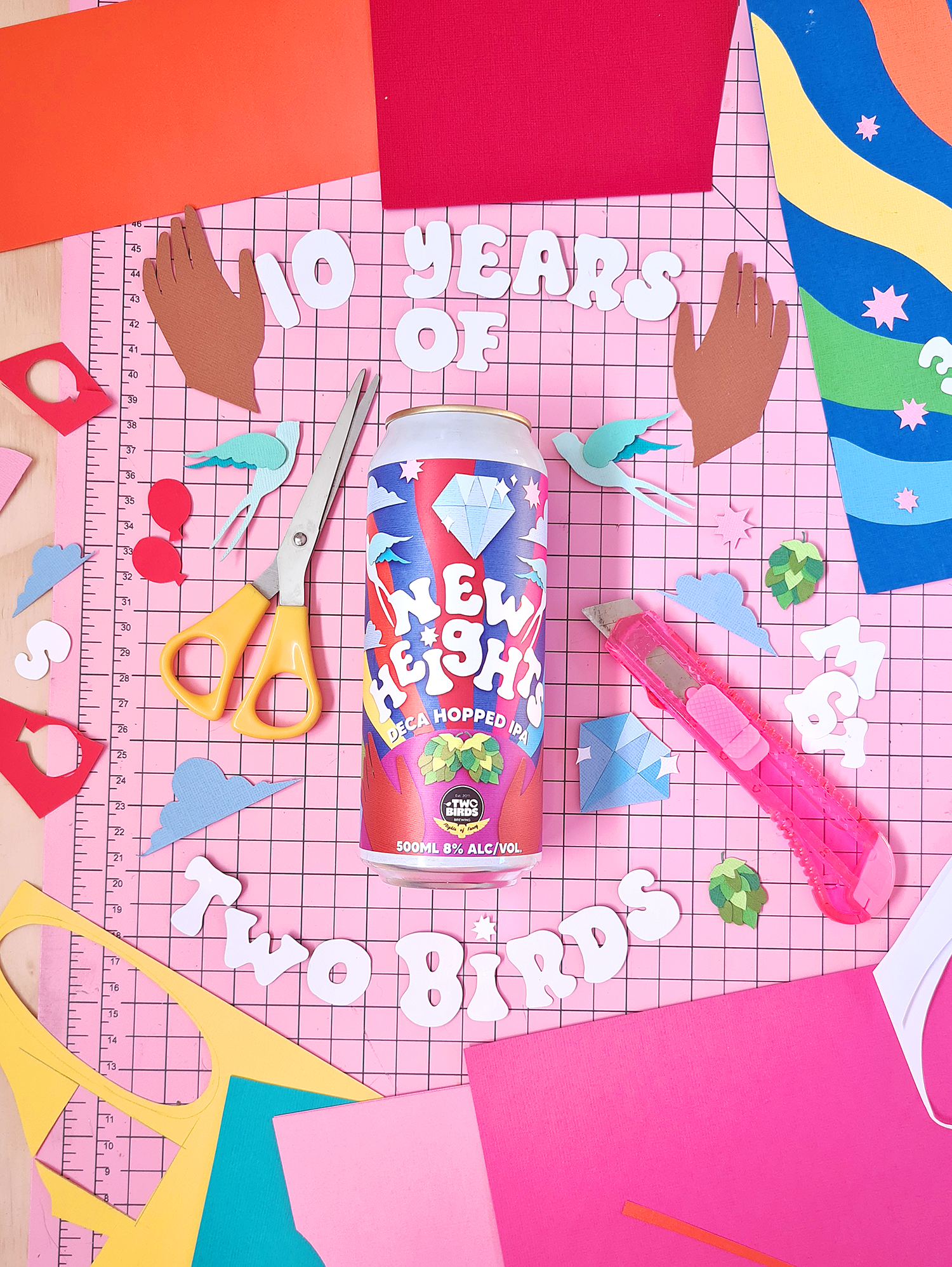 A colourful beer can with papercraft collage illustrated label lies on a studio desk surround by the text “10 years of Two Birds”, and various craft supplies. 