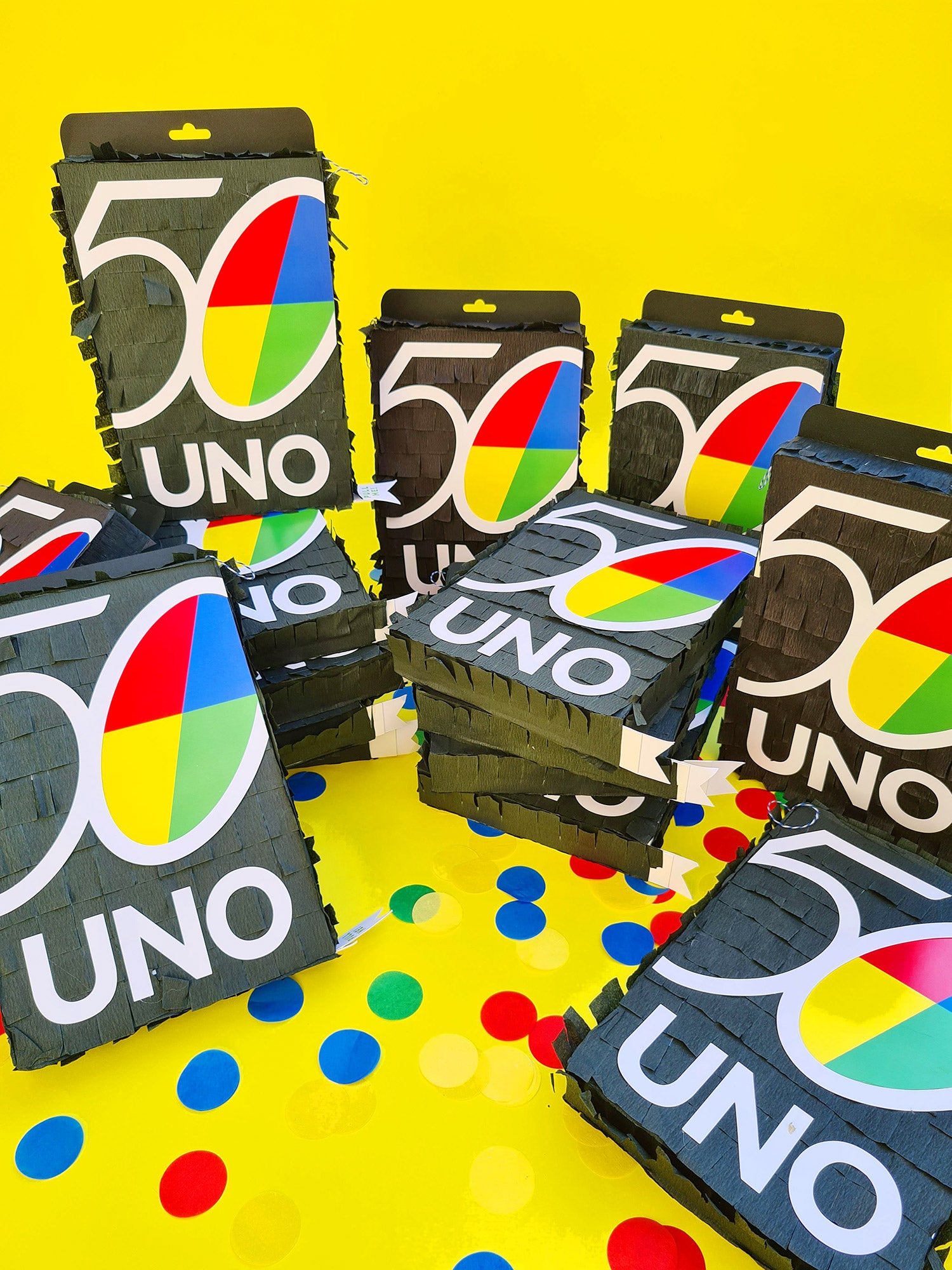 A collection of piñatas in the shape of black UNO playing card boxes, with the UNO logo on the front sit on a yellow background, surrounded by confetti in blue, yellow, red and green. 