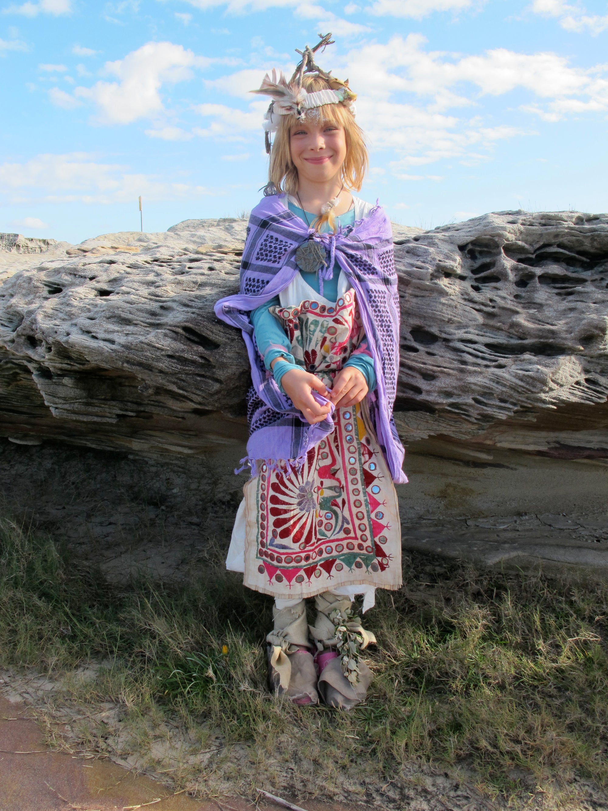 A young child dressed in a post-apocalyptic handmade costume stands against the rocks at a beach. 