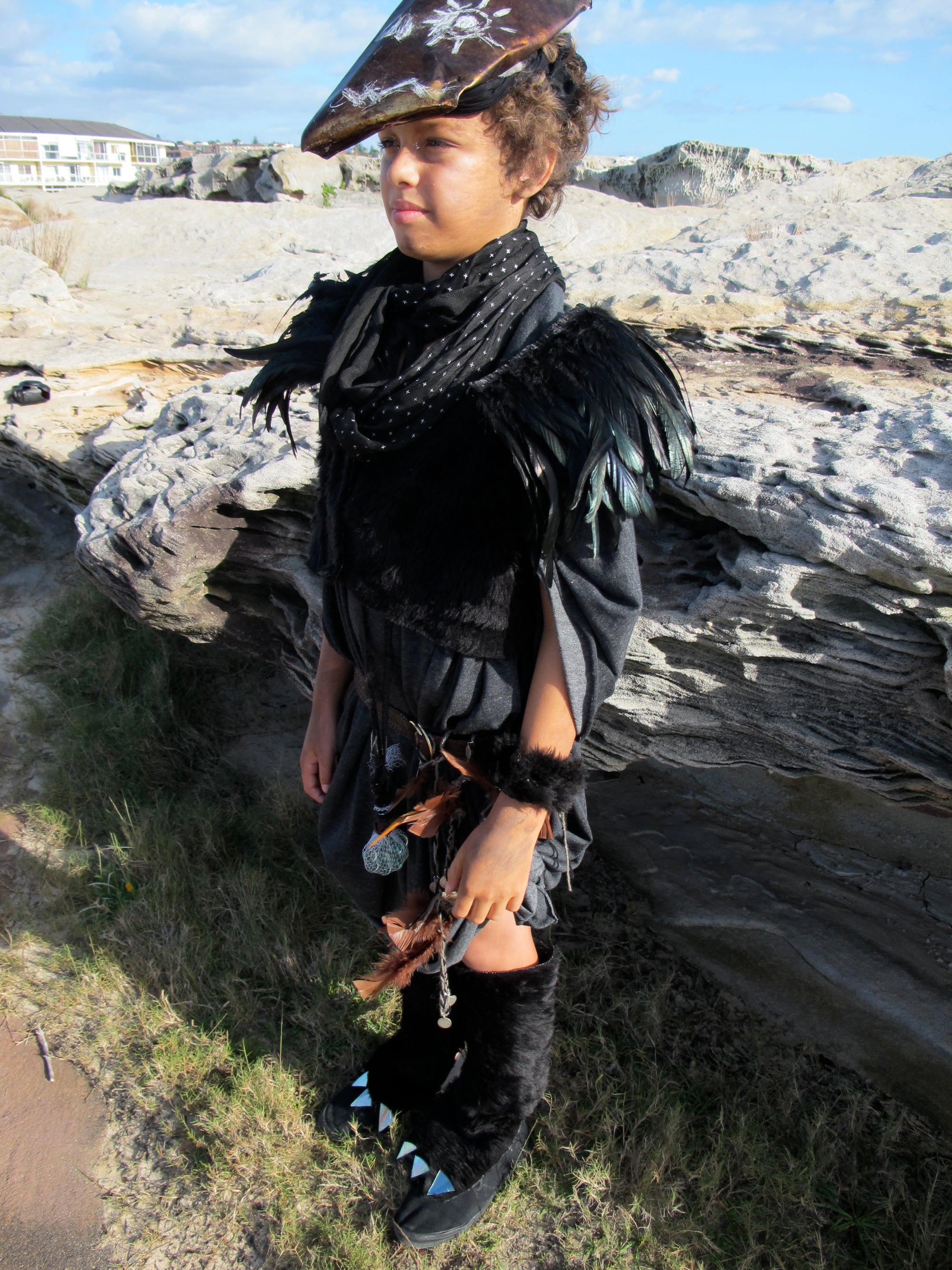 A young child dressed in a post-apocalyptic handmade costume stands against the rocks at a beach. 
