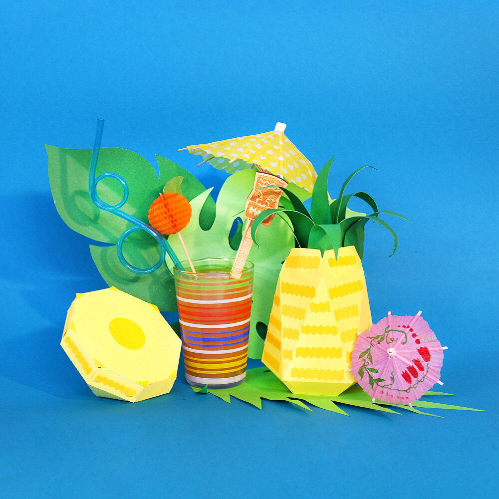 A tropical cocktail in a striped glass sits among a collection of tropical papercraft props, against a blue background.