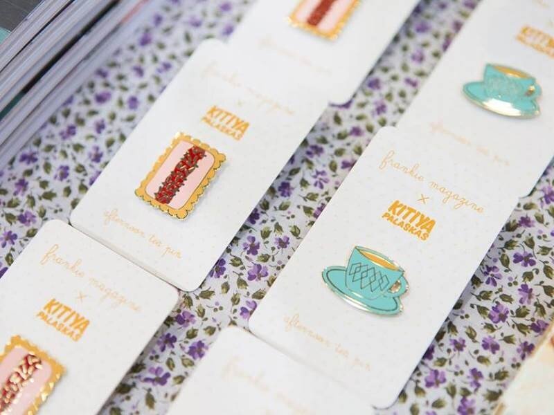 A collection of afternoon-tea enamel pins on printed packaging backing cards. One pin design is a cup of tea and one is a biscuit.