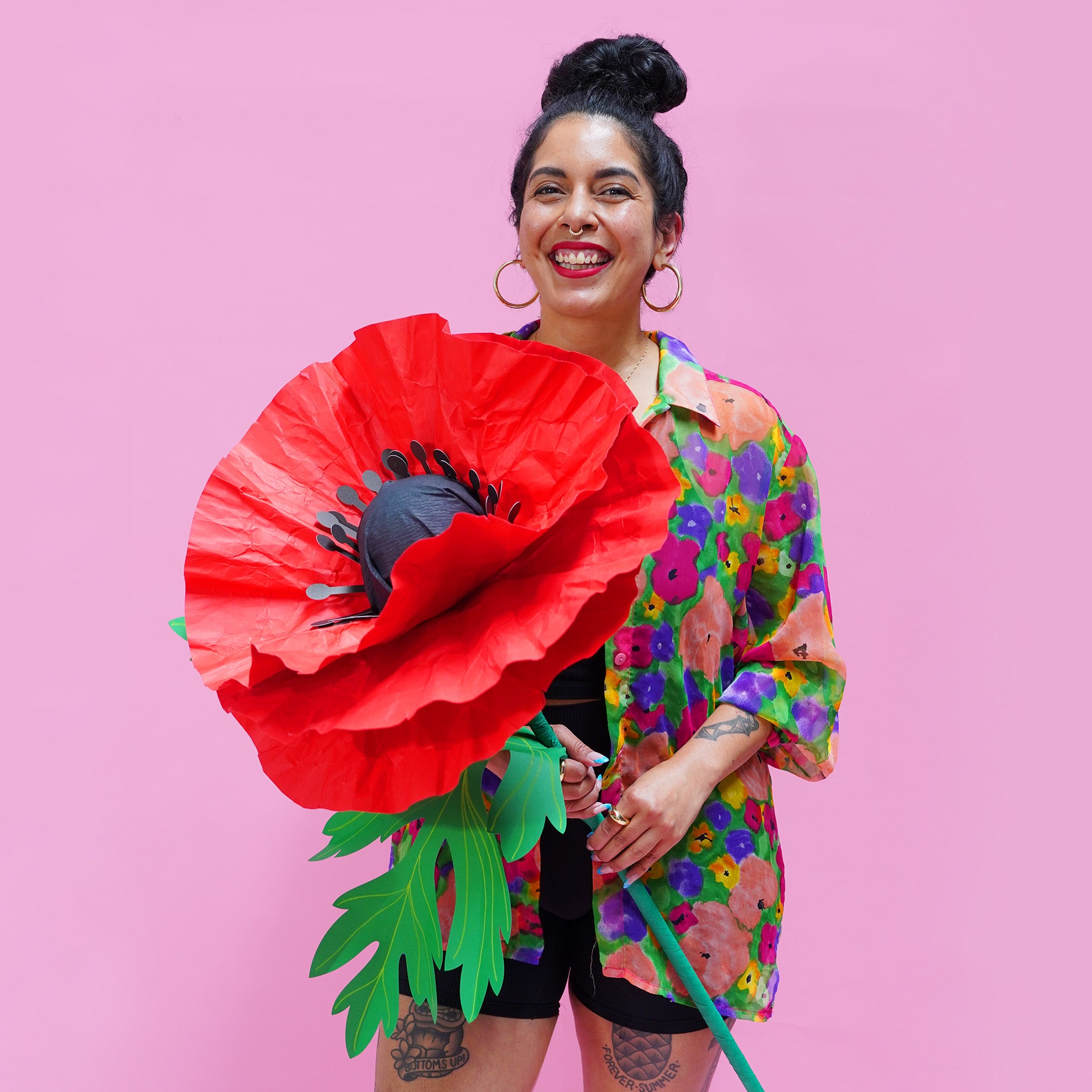 Kitiya, a woman with brown skin and black hair, holds a giant paper poppy prop in front of a pink background. She wears a colourful floral shirt and black shorts.