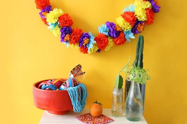 A vibrant and colourful handmade tissue paper flower garland hangs on a yellow wall. Below the garland is a stool with a bowl of accessories and some cactuses in glass bottles. 