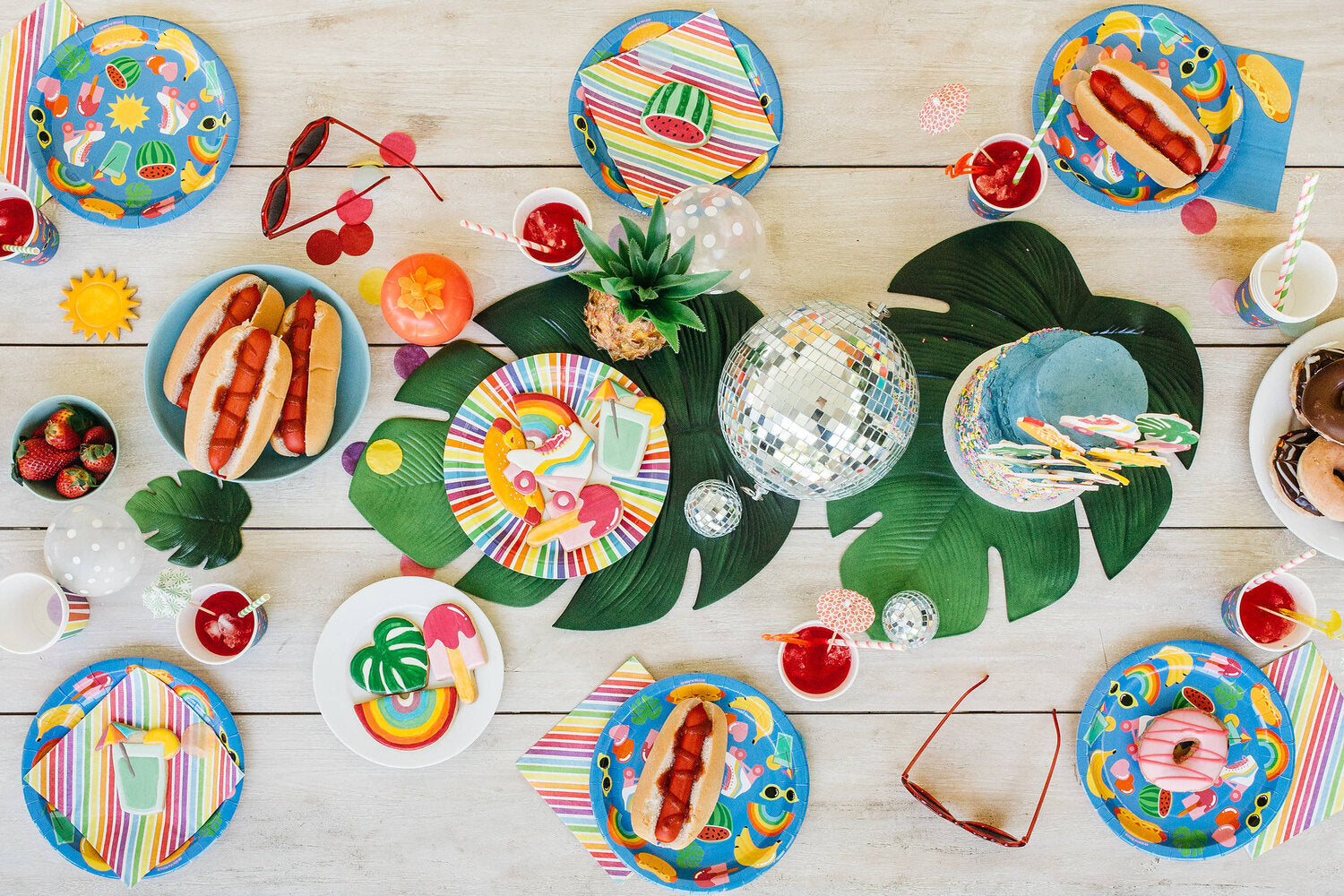 A collection of vibrant partyware in a party setting. The partyware includes plates, cups, party bags and piñatas and features a blue background, with tropical illustrated motifs. 