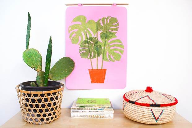 A pink wallhanging with block-printed green monsteral leaves in a brown pot. The wallhanging hangs on a white wall surrounded by books, a cactus and a desk.