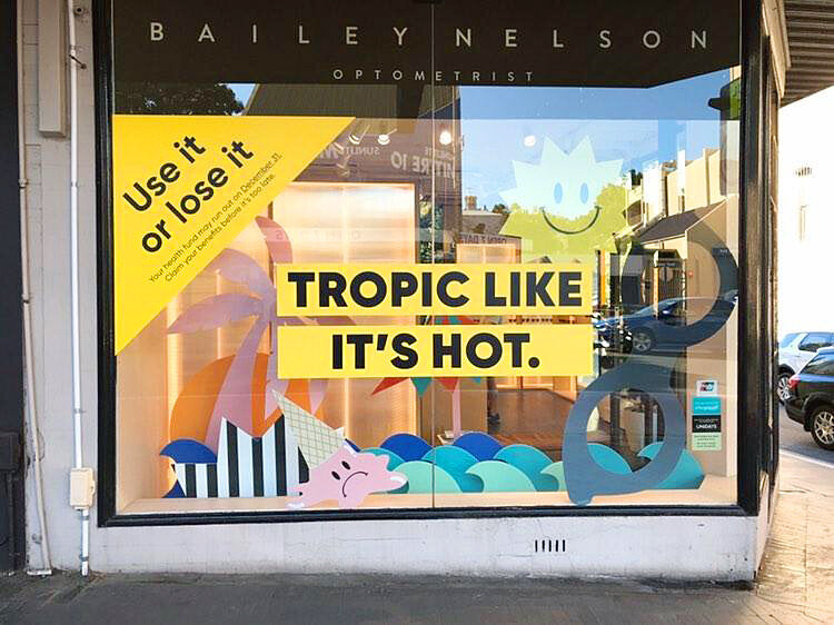 A colourful installation of cut-out shapes decorates. Bailey Nelson retail store window. The  window props include a giant pair of eyeglasses, stripy beach umbrella, sun with smiley face, black and white striped starburst shape, melted ice cream with sad 